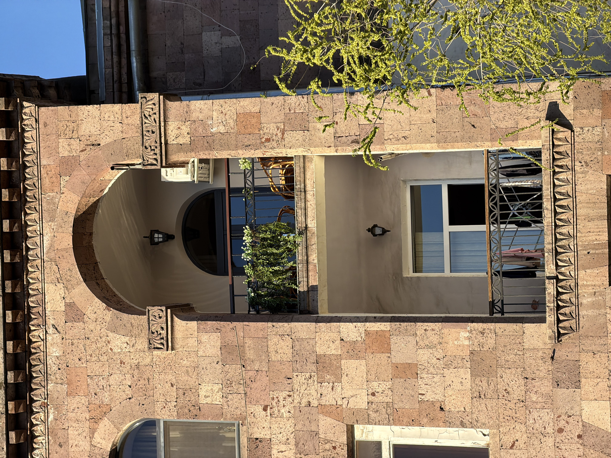 The image displays a detailed view of a building facade, characteristic of Yerevan, Armenia, constructed from variegated stone blocks, likely tuff, ranging in color from reddish-brown to lighter shades. The central and dominant feature is a large, deep arched opening, within which a recessed area or balcony is visible.
To the left of the main arch, a vertical decorative element with a repeating pattern adorns the stone facade. Inside the main arch, the recessed space features a rounded arch on its left side, creating a deep alcove. The interior walls of this recessed area are a lighter, beige color, and two wall-mounted light fixtures are affixed to them.
On the right side of the recessed space, a rectangular window with white frames is prominent, set behind a decorative metal railing. Through the window, white curtains or blinds are visible, and the glass reflects parts of the sky and surroundings. A wicker chair and several potted plants, one bearing small white flowers, are discernible behind the railing, indicating a functional outdoor space.
Above the building, in the upper right portion of the image, the branches of a tree with vibrant, light green leaves or blossoms extend into the frame, casting dappled shadows on the stone. A clear blue sky is visible in the top left corner, confirming sunny weather conditions. Below the main arched opening, two other windows are partially visible at the bottom of the frame. The strong sunlight illuminates the scene, highlighting the textures of the stone and architectural details. No text is visible in the image.