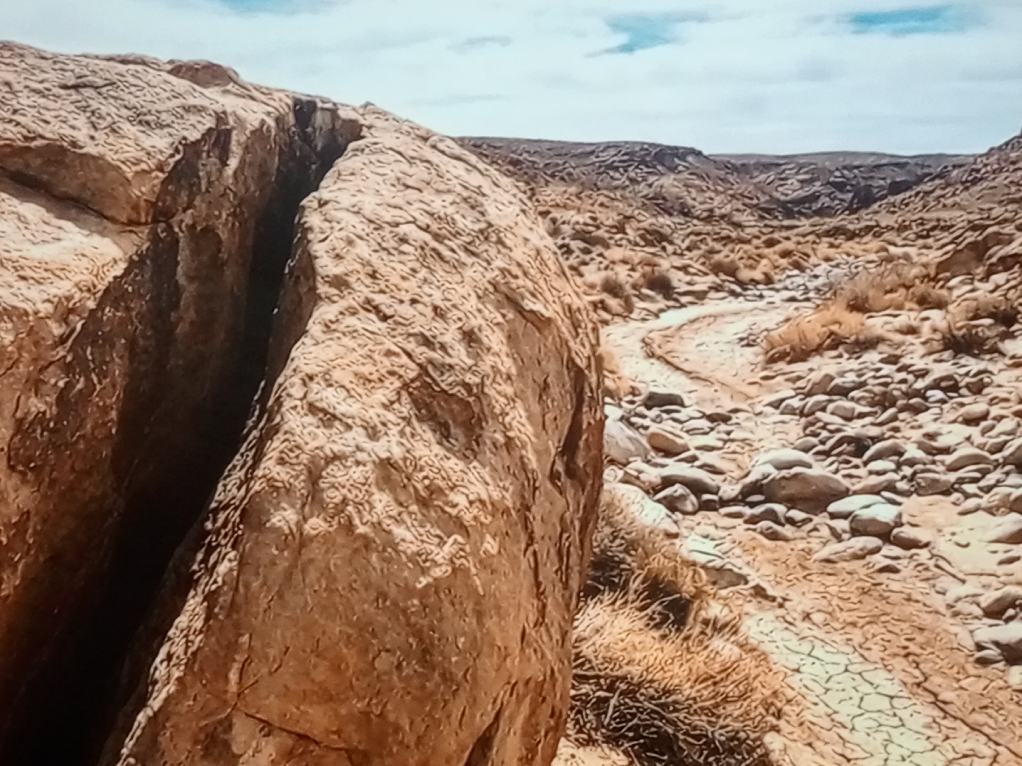 A rocky, arid canyon landscape under a partly cloudy sky. The foreground is dominated by a large, weathered boulder with a deep fissure running through its center. To the right, a dry, winding path made of pebbles and sand leads into the distance, flanked by sparse, dry vegetation and more rocky terrain. The background features rolling hills and distant mountains, all rendered in muted earth tones. The scene appears to be a natural, undeveloped area, likely in a desert region. There are no people or specific urban elements visible.