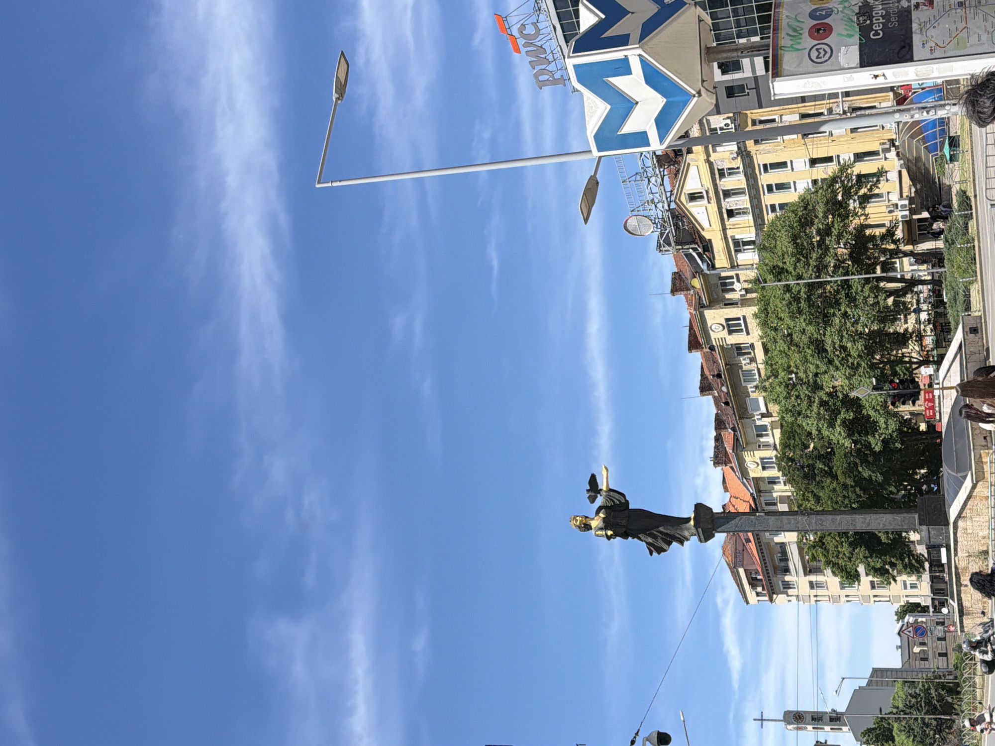 The image depicts a cityscape under a bright blue sky with wispy clouds. A statue stands prominently in the mid-ground, depicting a figure holding an object, possibly a wreath or a branch. Behind the statue are buildings of various heights and architectural styles, suggesting a historical or urban setting. To the left of the statue, there is a streetlight pole. At the top, there is a blue and white sign with the name of a company "PwC" above it, as well as a sign for a metro station. People are visible in the background.