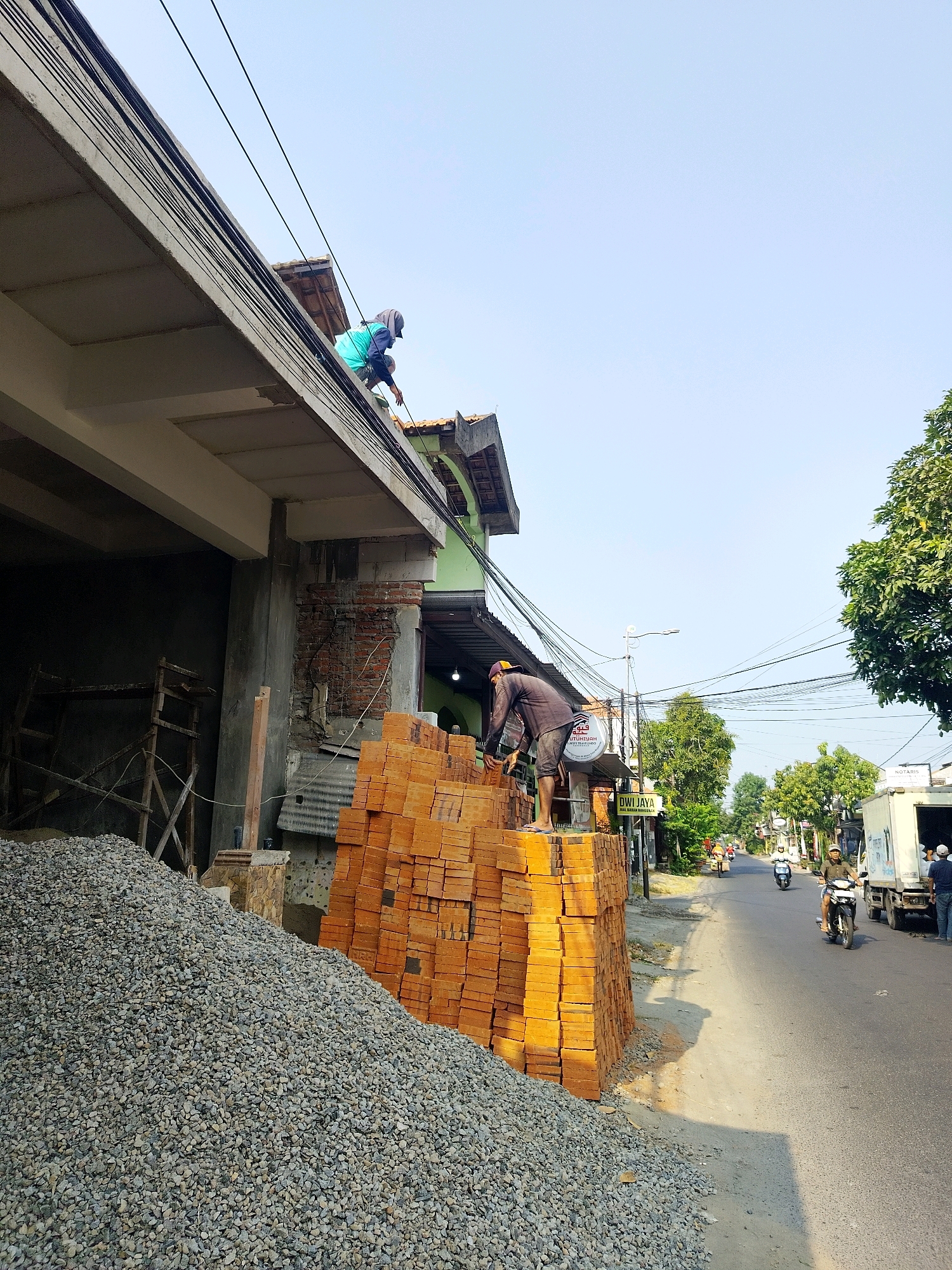 The image depicts a street scene in Mojokerto, Indonesia, with a focus on a construction site. In the foreground, a large pile of gravel is visible next to a stack of orange bricks. Two construction workers are present. One worker, wearing a teal shirt and a head covering, is positioned on the roof of a building, seemingly working on electrical wires. Another worker, wearing a dark shirt and shorts, is standing on top of the brick pile, appearing to be engaged in some task related to the construction materials.
The building under construction features exposed brickwork and a concrete overhang. Power lines crisscross the sky above the street. Further down the road, other buildings and trees line the street, and several motorcycles and a truck are visible, suggesting typical street activity. The overall atmosphere is that of a sunny day in a developing urban area.