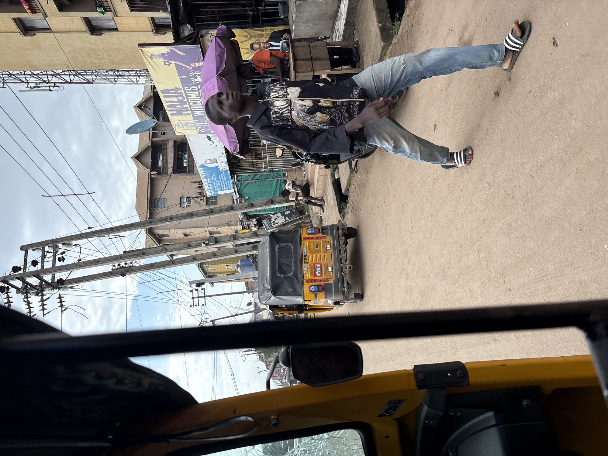The image captures a street scene in Nkpor, Nigeria, viewed from the interior of a yellow auto-rickshaw. In the foreground, the side and part of the windshield of the auto-rickshaw are visible, framing the rest of the scene. A young Black male is walking from right to left in the mid-ground, wearing a black zip-up jacket with a graphic design and light blue distressed jeans. He has on black and white striped slide sandals. Behind him, a yellow auto-rickshaw with a black canvas top is parked on the dirt road. Several multi-story buildings line the street, some with balconies and windows. Overhead, a complex network of utility poles and wires is visible against a cloudy sky. A large banner with the text "NAIJA WELCOMIS" is displayed on the side of one of the buildings. To the right of the walking figure, a table with a purple umbrella provides shade, and two men are visible behind it, one in an orange shirt and another in a suit. The overall environment suggests a bustling urban setting with everyday activities taking place.