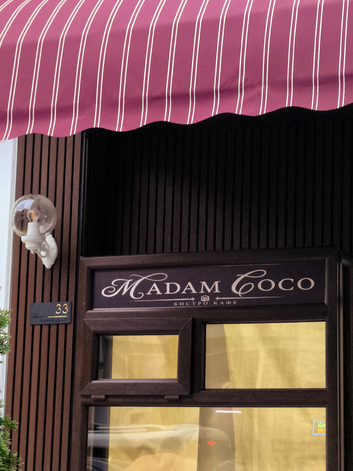 The image shows the storefront of a cafe named "MADAM COCO". The cafe has a sign with this name written in elegant script. Below the name, there is text in Cyrillic script, which reads "БИСТРО КАФЕ" (Bistro Cafe). The storefront features a dark brown frame with two windows reflecting a yellow light. A number "33" is displayed on a dark plaque to the left of the entrance. Above the entrance, there is a striped awning in dark red and white. A round glass light fixture is mounted on the wall to the left of the sign. The building facade is made of dark brown vertical wooden planks. This storefront is located in Batumi, Georgia.