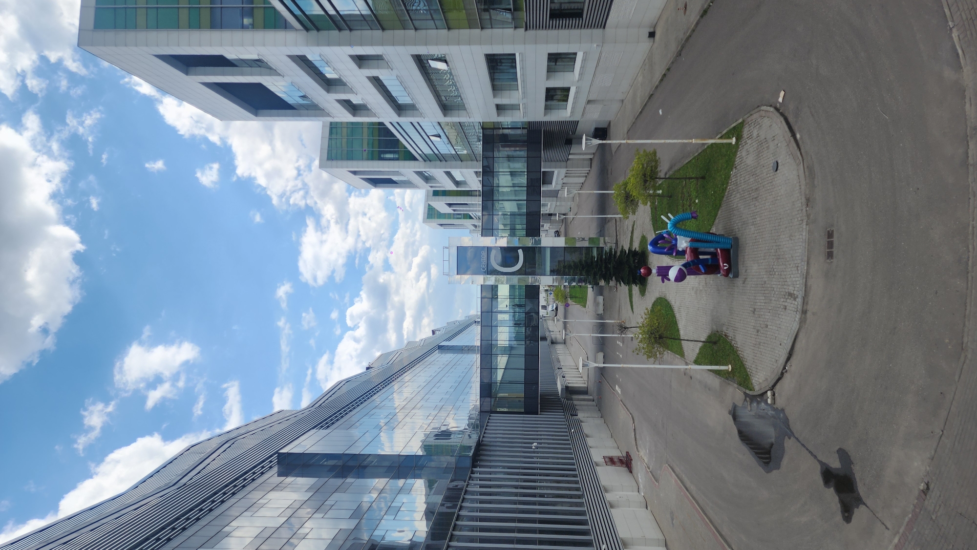 The image shows a modern architectural landscape, likely in Moscow, Russia. The scene features a mix of glass and concrete buildings under a blue sky with scattered clouds. A prominent glass building with a cascading design is on the lower left, connected to a white, rectangular building on the upper left by a glass bridge with the letter "C" on it. A roundabout with landscaping, including trees and a sculpture, is to the right of the bridge. The ground is primarily paved, with some grassy areas near the roundabout. A puddle on the road shows a reflection of a person walking down the street.