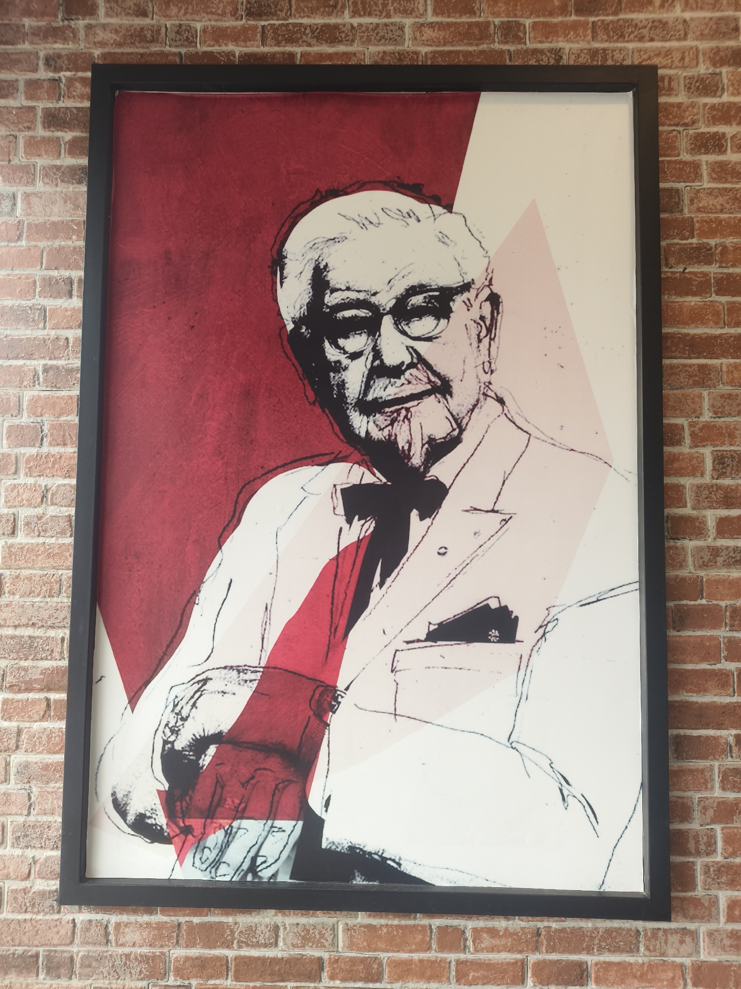 A framed portrait of Colonel Sanders, the founder of KFC, is mounted on a red brick wall. The portrait is in a pop-art style, with bold black lines and areas of red and white. Colonel Sanders is depicted wearing his signature white suit and black bowtie, with glasses and a goatee. His hands are visible in the foreground, with one resting on his chest. The background of the portrait is a abstract design with red and white geometric shapes. The wall it is mounted on is made of red brick with visible mortar. The image appears to be taken indoors, likely in a KFC restaurant. The location is Denpasar, Indonesia.
