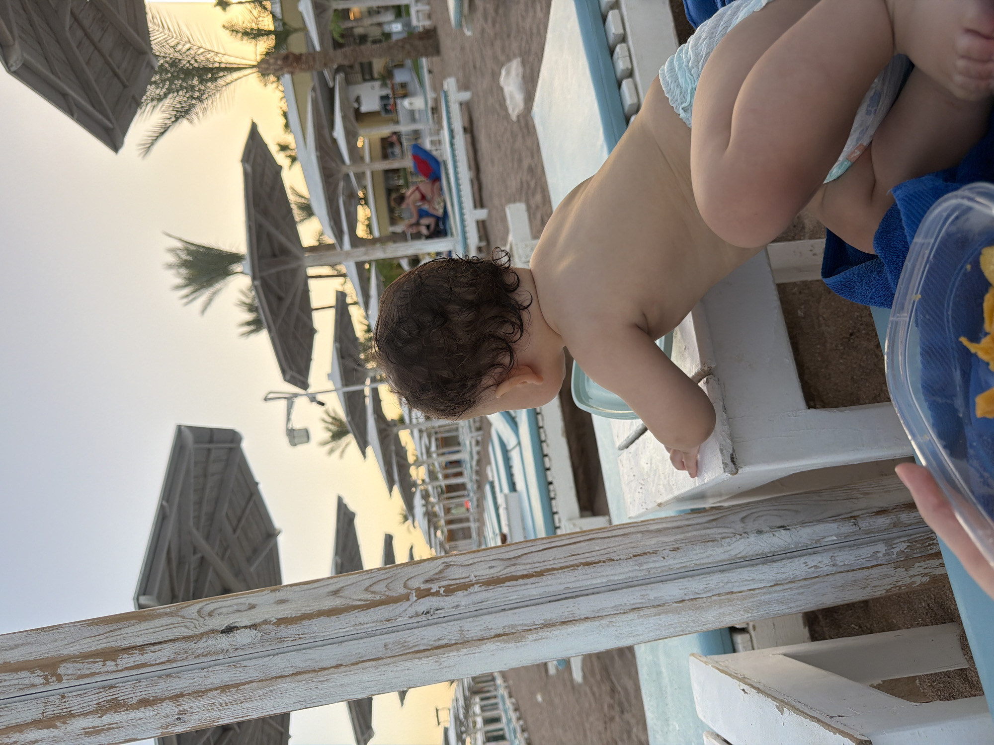 The image depicts a baby in a diaper leaning over a white wooden railing on a beach. The baby has curly brown hair and is reaching out with their hands. Behind the baby, a beach setting is visible with lounge chairs, umbrellas, palm trees, and people in the distance. The scene appears to be from a resort or hotel on a sandy beach, likely in a warm climate given the attire and setting. The lighting suggests it is either early morning or late afternoon, with the sun low on the horizon, casting a warm glow. The overall mood is relaxed and peaceful, characteristic of a vacation or a day at the beach.