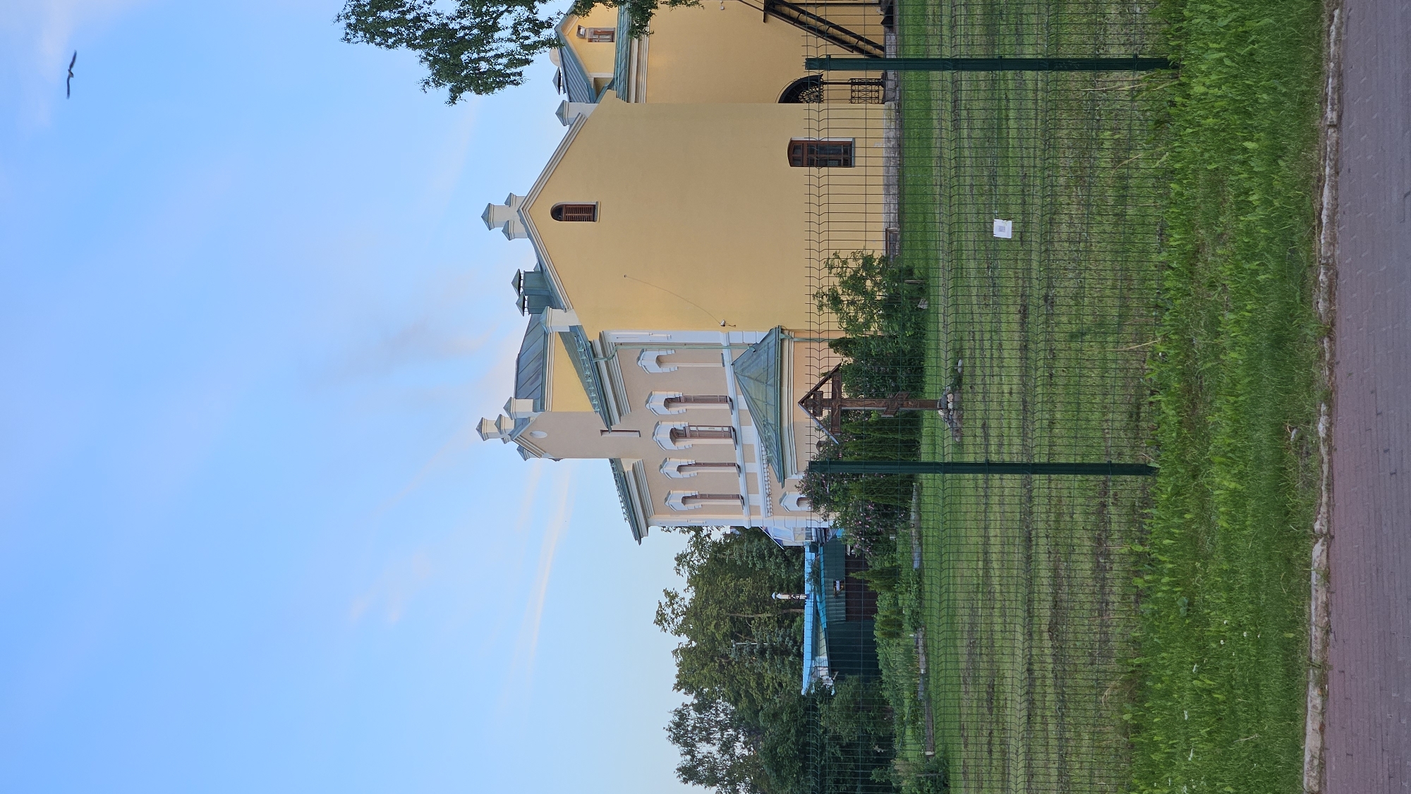 The image captures a yellow building with a green roof and trim, situated in an outdoor setting. A dark green metal fence runs across the foreground, partially obscuring a grassy area and a patch of red brick pavement. Behind the fence, the building is flanked by trees and other greenery. A wooden cross stands in the yard in front of the building, and a small white sign is attached to the fence. The sky above is a pale blue with some wispy clouds, and a bird is visible in flight. The overall scene suggests a tranquil, possibly religious or historical, location.