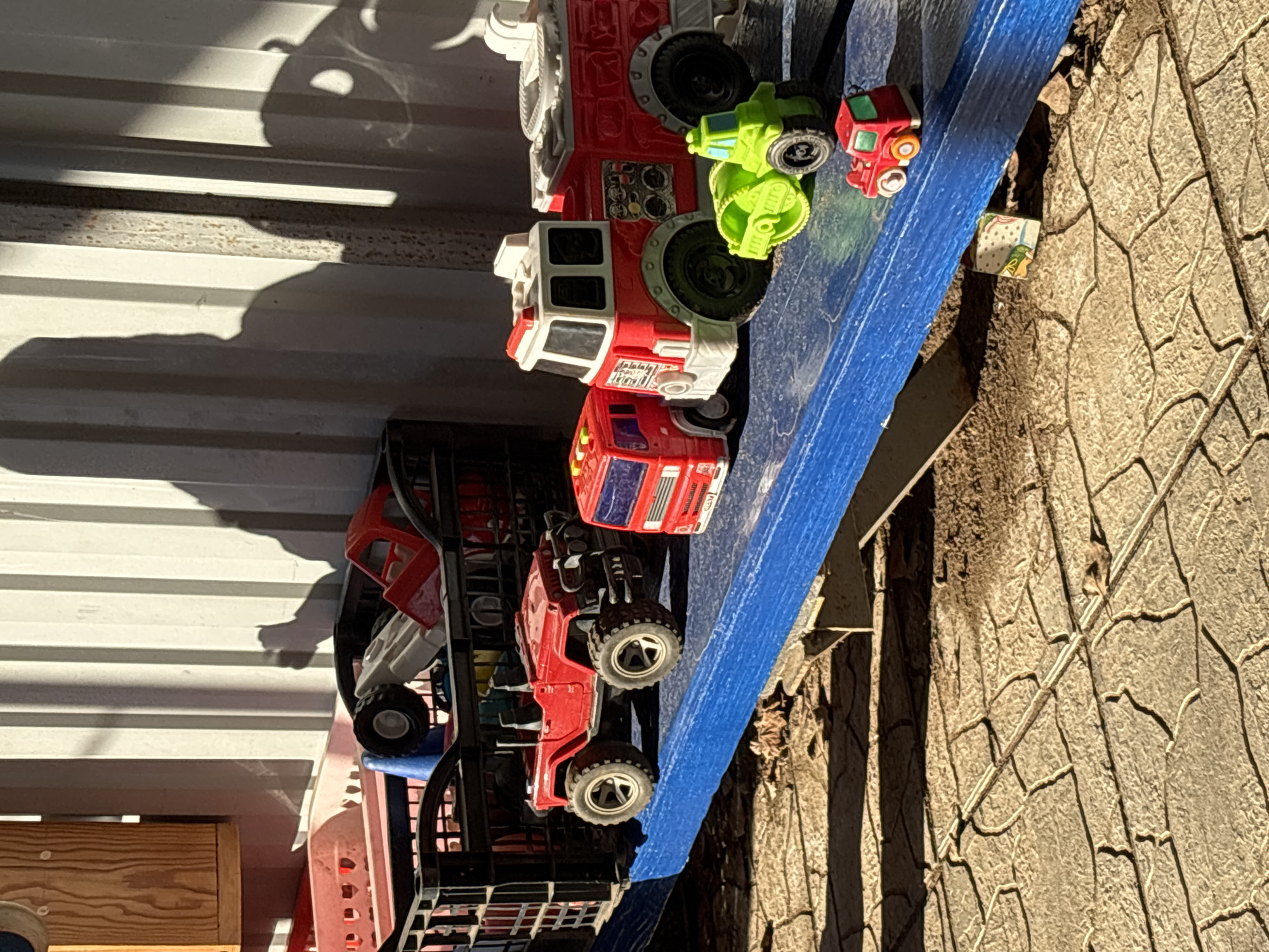 The image depicts a collection of toy vehicles, including fire trucks and other construction vehicles, arranged on a blue surface. The setting appears to be outdoors, possibly near a textured wall or surface. There is also a corrugated metal structure in the background, casting shadows. The location in the city of Chaikovsky, Russia is not determinable from the image.