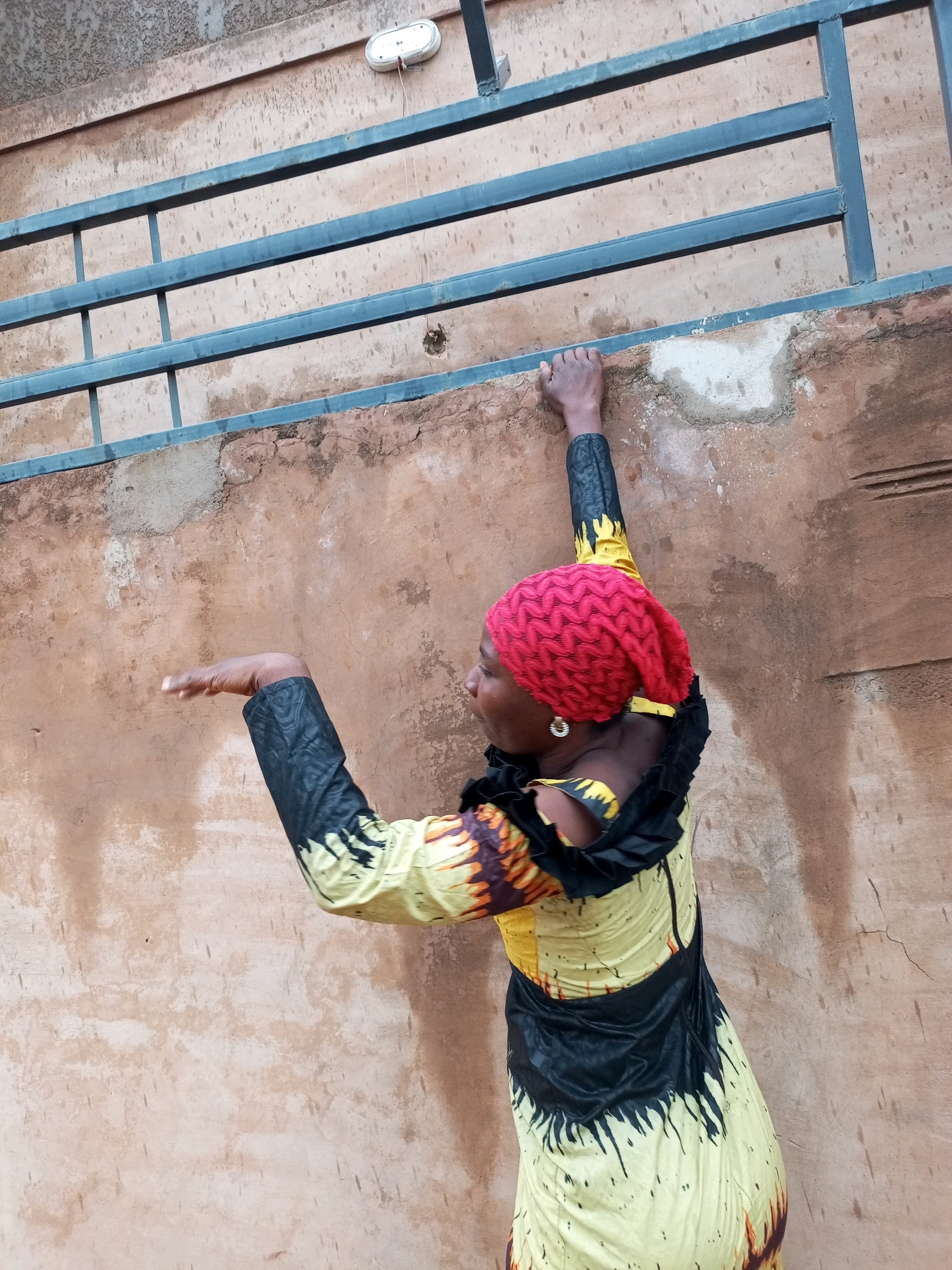A Black woman is pictured outdoors, wearing a red knitted headscarf and a yellow and black patterned dress. Her left arm is raised, resting on a horizontal metal bar that is attached to a textured, light brown wall. The wall appears weathered, with some discoloration and visible cracks. Her right arm is bent and extended outwards. The background consists of the wall and the metal bar, with a small white fixture visible at the top of the wall. The scene is consistent with an outdoor location in the city of Tempure, Nigeria, likely an exterior wall of a building.