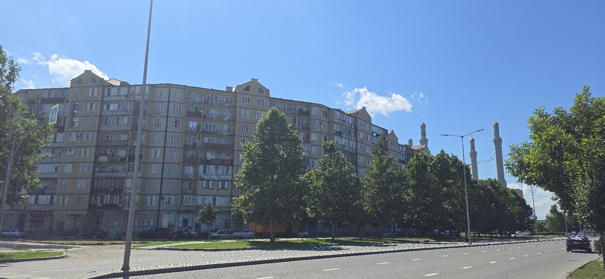 This is a horizontal, eye-level outdoor shot of a city street in Grozny, Russia, on a bright, sunny day. The sky is a clear, vibrant blue with a few wispy white clouds. The foreground is dominated by a paved road with white lane markings. To the left of the road is a strip of green grass. Behind the grass, a large, multi-story apartment building with a beige facade occupies the middle ground. The building has many windows and balconies, and a distinct gabled roofline. Trees with lush green foliage line the street in front of and beside the apartment building, partially obscuring it. In the distance, several tall, slender towers with golden domes are visible, hinting at a mosque or religious complex. A black car is driving on the road in the right-hand portion of the image. A yellow pedestrian crossing sign is visible on a pole near the apartment building. The overall impression is one of a typical urban scene on a pleasant day. No people are clearly discernible in the image. There is some faint text visible on the building facades, including "BIORISE" and "THERAPY."