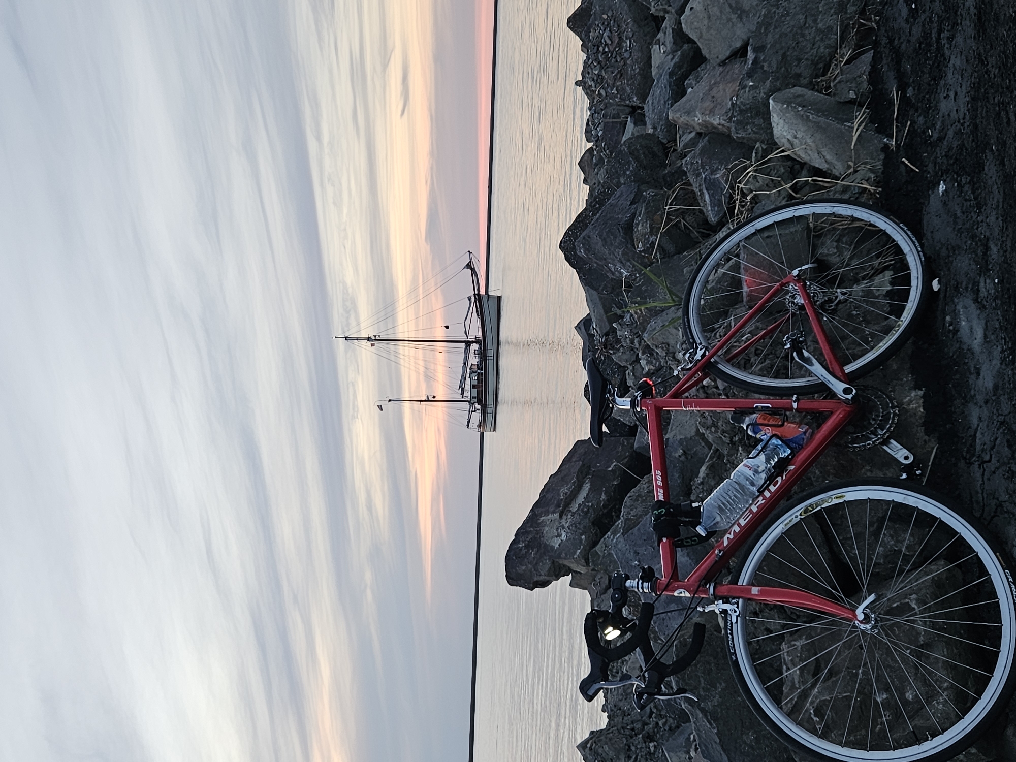A red Merida ME 905 bicycle is parked on a rocky shore, facing the water. The bicycle is positioned in the foreground, with its front wheel on the left and rear wheel on the right. A water bottle is secured in a cage on the frame. In the background, a sailboat with two masts is sailing on the calm water, under a sky filled with wispy clouds. The scene is set at the Oostvaardersdiep in Lelystad, Netherlands, at dusk. The water reflects the soft colors of the sunset. The rocky shore appears to be a breakwater or a path along the water.