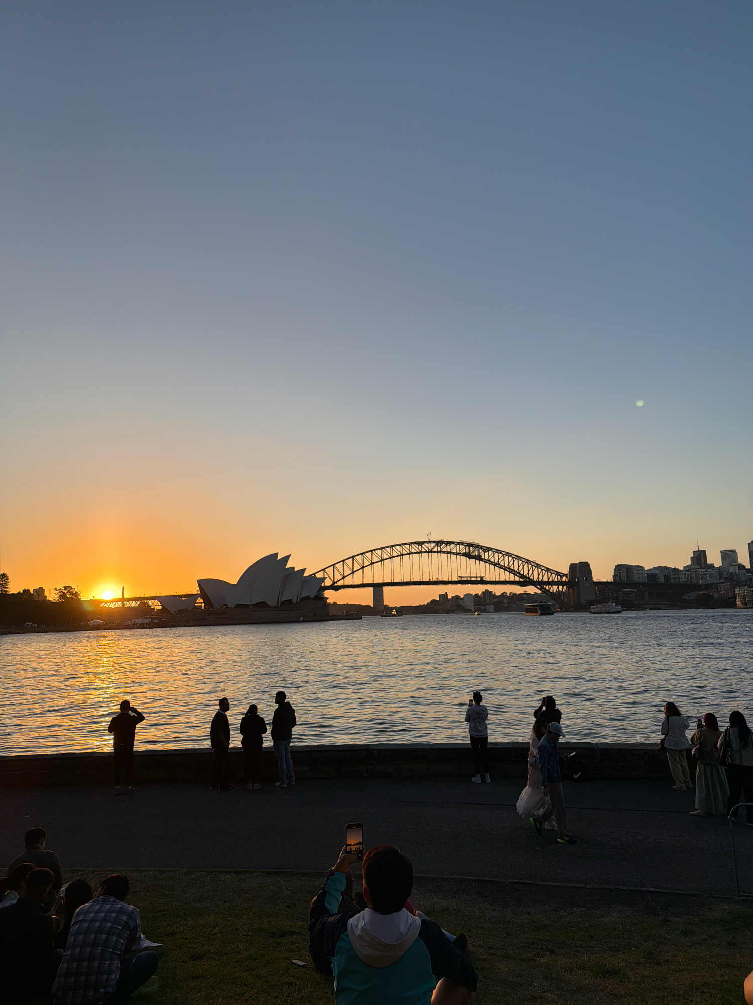 The image captures a sunset over Sydney Harbour in Australia. In the foreground, a group of people are seated on the grass, with one individual holding up a phone to take a picture. Beyond them, a paved walkway lines the harbor, where several silhouettes of people are standing and observing the sunset. The water of the harbor reflects the warm orange and yellow hues of the setting sun. Dominating the background are iconic Sydney landmarks: the Sydney Opera House with its distinctive white sails, and the Sydney Harbour Bridge, a massive steel arch structure. The sky is a gradient of pale blue to soft orange. Boats are visible on the water, and the city skyline with its buildings can be seen in the distance. The overall scene depicts a serene evening at a popular tourist spot.