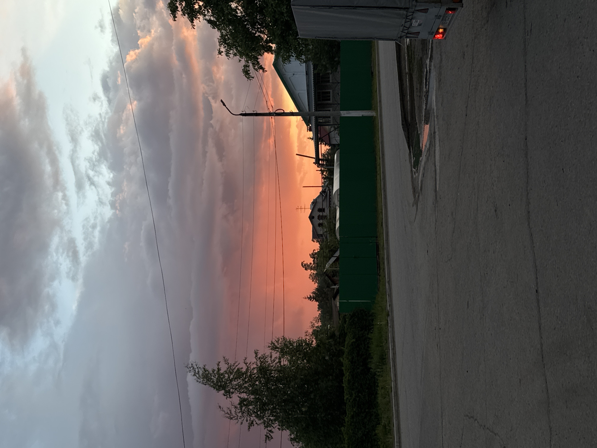 A medium-shot, vertical photograph taken in the city of Noviy, Russia, depicts a street scene at sunset. The sky is a vibrant mix of pink and gray clouds. A road is visible on the right side of the frame, with a green fence running parallel to it. Trees line the bottom of the image. A utility pole and wires intersect the sunset. The back of a semi-trailer is present at the top of the image. Buildings are visible in the distance between the green fence and the setting sun.