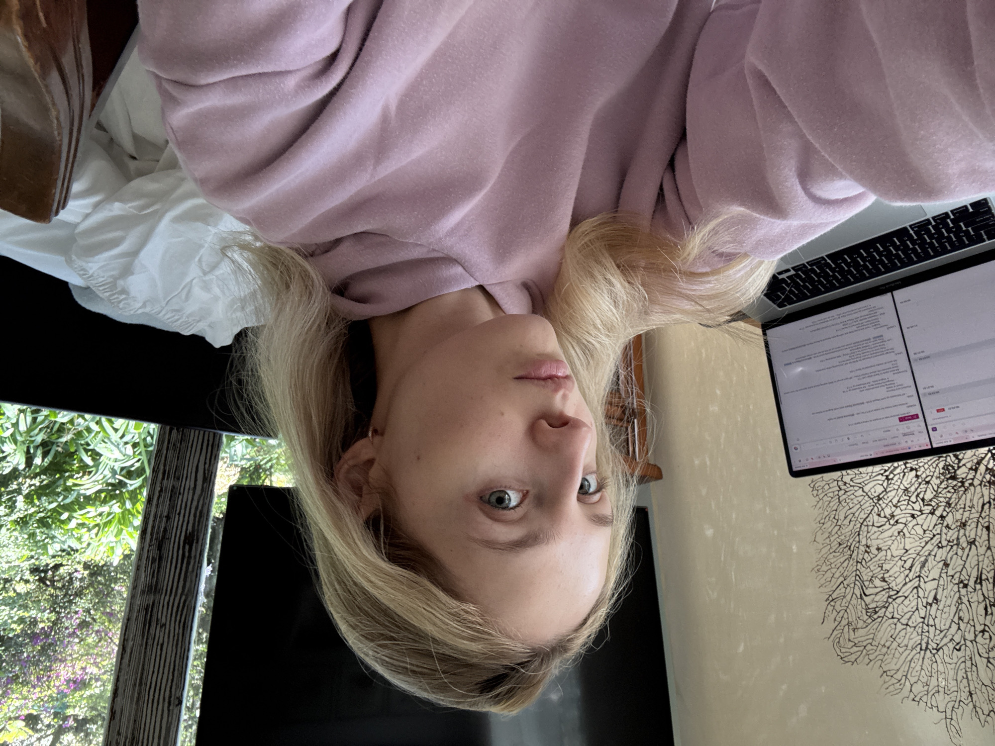 In an indoor scene, a young, fair-skinned woman with blonde hair pulled back and a pink hooded sweatshirt is captured in a selfie. She is seated in front of an open laptop displaying text and graphics. To her left, a decorative, coral-like object adorns a light-colored wall. In the background, a television screen is visible, along with a wooden chair and a window revealing lush green foliage and purple flowers outside. The setting appears to be a home environment. The location is not identifiable as Ciudad de México, Mexico, or any specific city.