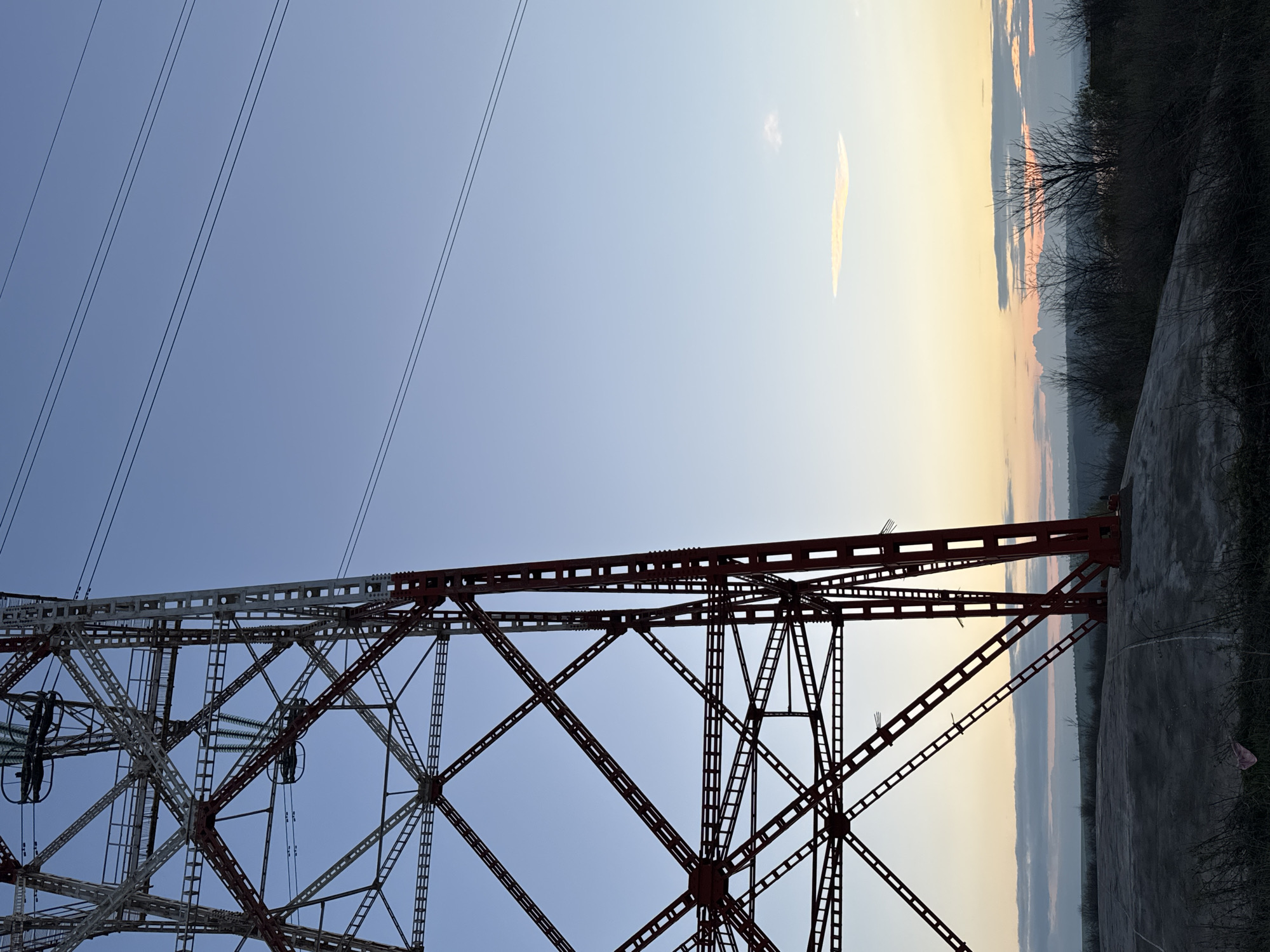The image shows a high-voltage electricity pylon near Kozloduy, Bulgaria, with a segment of the sky at sunset in the background. The pylon has a red and white lattice structure. Overhead power lines stretch to the left of the pylon. The land and some vegetation are visible on the right side of the image, below the sunset.