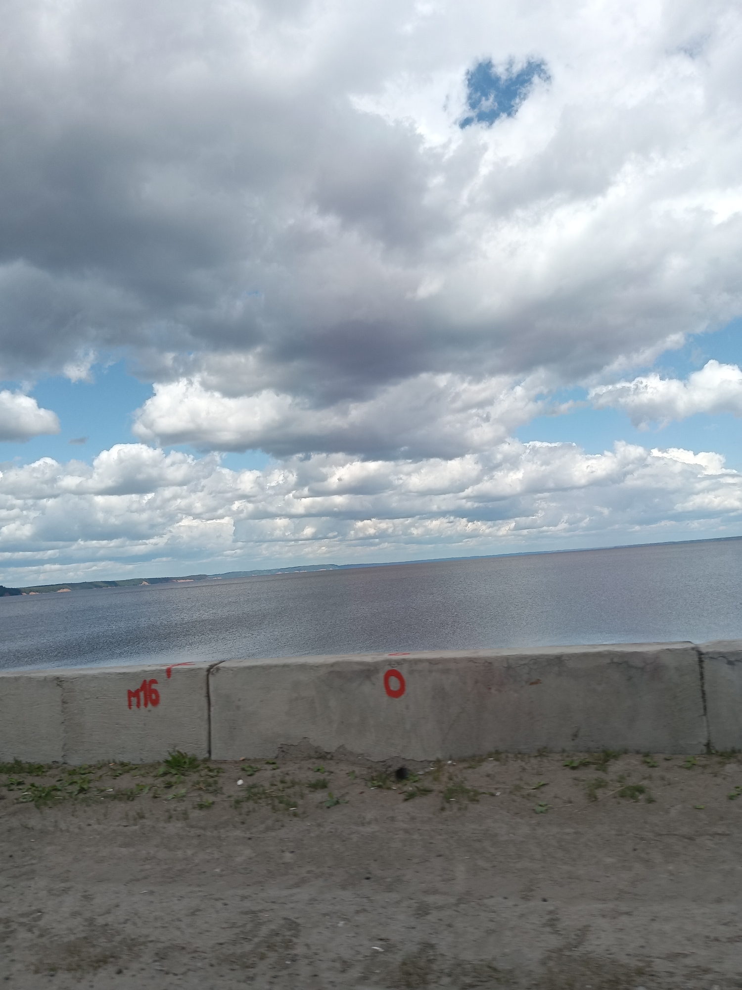 The image depicts a waterscape scene under a cloudy sky. In the background, a body of water stretches toward the horizon, meeting the distant shoreline. The sky is filled with numerous grey and white clouds. In the foreground, there is a concrete barrier. Red graffiti is on the concrete barrier. Beyond the barrier, there is a dirt ground with scattered vegetation.
The city of Chaikovsky, Russia is potentially the location due to the presence of M16 and the overall landscape.