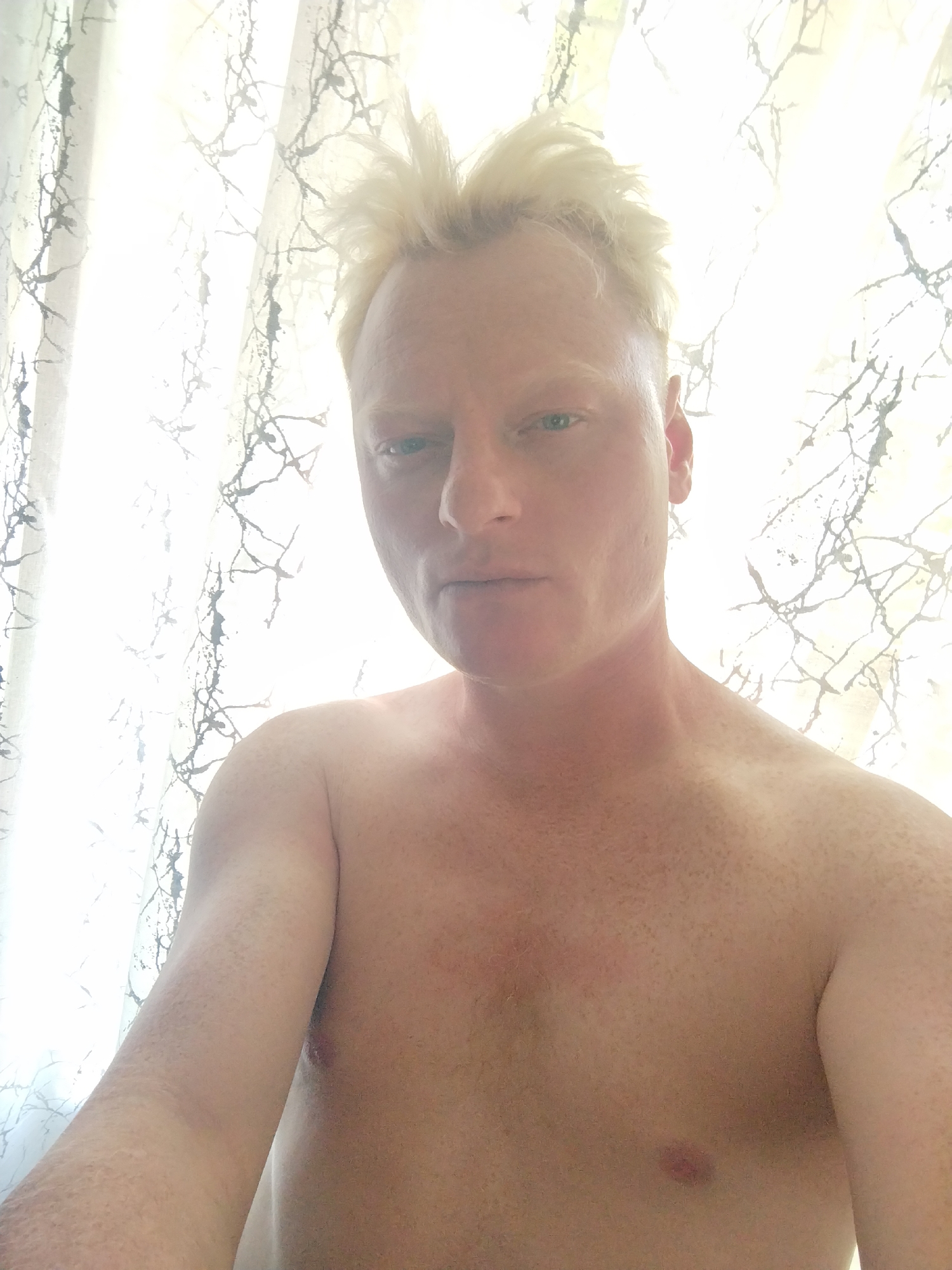 The media file is a selfie of a fair-skinned man with short, spiky blonde hair. He is shirtless, revealing his upper chest and shoulder which are dotted with freckles. He has blue eyes and is looking directly at the camera with a neutral expression. The background consists of a sheer curtain with a dark, tree-like pattern. The lighting is bright, suggesting it is daytime and natural light is coming from behind the curtain. The overall scene appears to be indoors, possibly in a bedroom or living area. There is no visible text in the image.