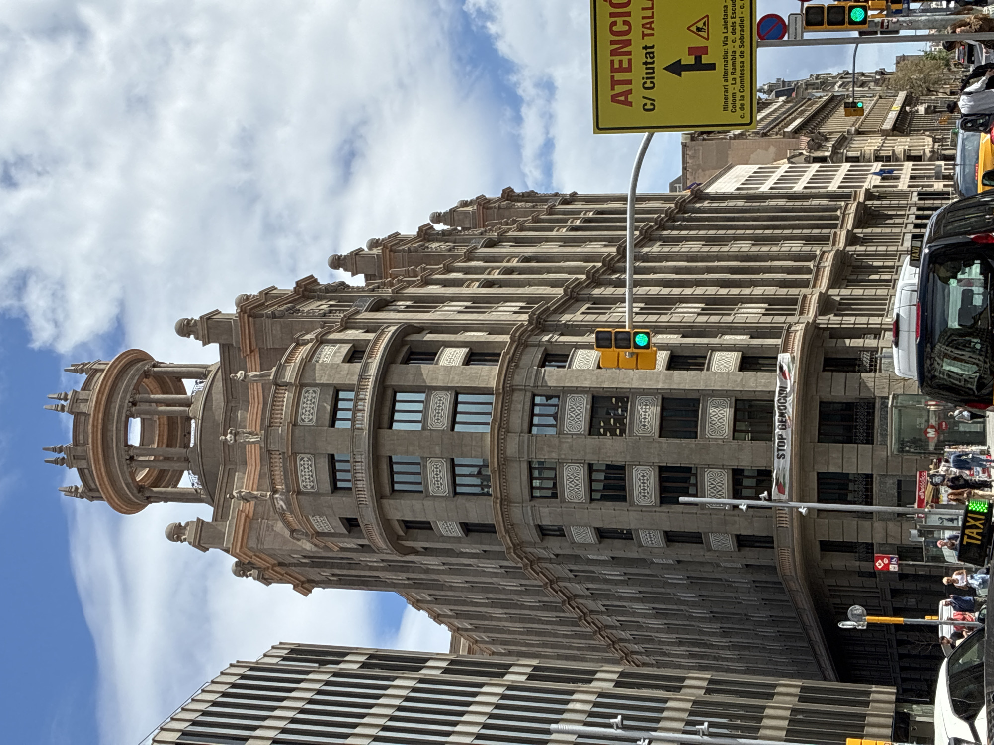 The image shows a tall, ornate building with a domed top in Barcelona, Spain. The building has a light grey facade with rows of windows and decorative detailing. A street sign that reads "ATENCIÓ C/ Ciutat TALLA" and the words "STOP GENOCIDI" are present. There are traffic lights with a green light showing. The street has pedestrians, a taxi, and other cars. The sky is partly cloudy. A modern striped building is visible at the bottom left.