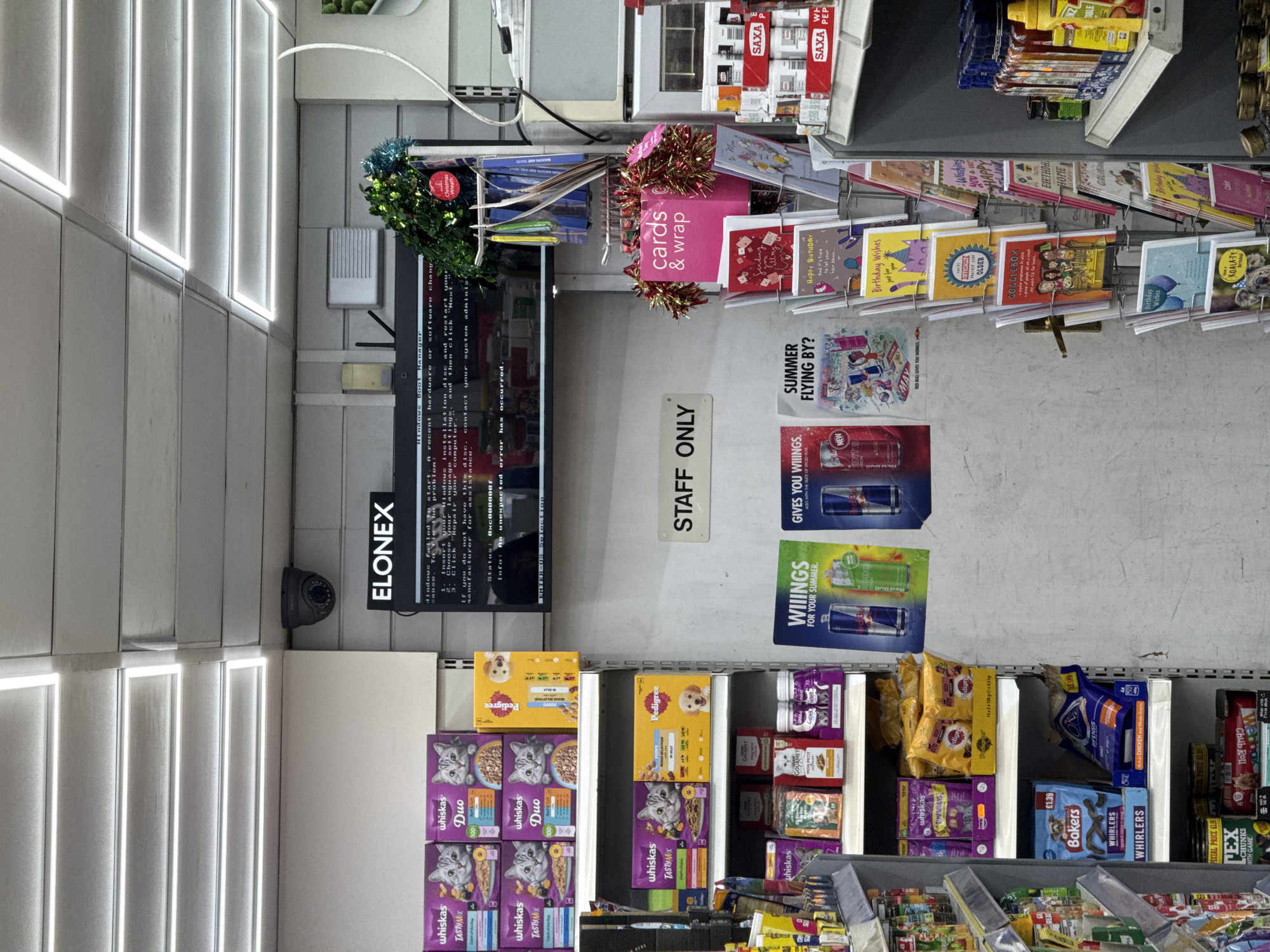The image shows the interior of a store, potentially in London, United Kingdom, given the presence of British product brands and retail displays. Shelves are stocked with pet food, particularly cat food brands like Whiskas and Gourmet, and dog food from Pedigree and Bakers. Adjacent shelves feature greeting cards, including birthday cards, and promotional displays for Red Bull.
A digital display, labeled "ELONEX," shows a Windows Boot Manager error message, suggesting a computer malfunction. The store environment is illuminated by linear overhead lights. A security camera is mounted near the digital display.
A sign reads "STAFF ONLY." Christmas-themed decorations are present above the greeting cards, including a small wreath and tinsel around a "cards & wrap" display.
The shelves are also stocked with spices, featuring SAXA salt. There are no people visible in the image. The overall impression is that of a typical retail store environment with a variety of products available for sale.