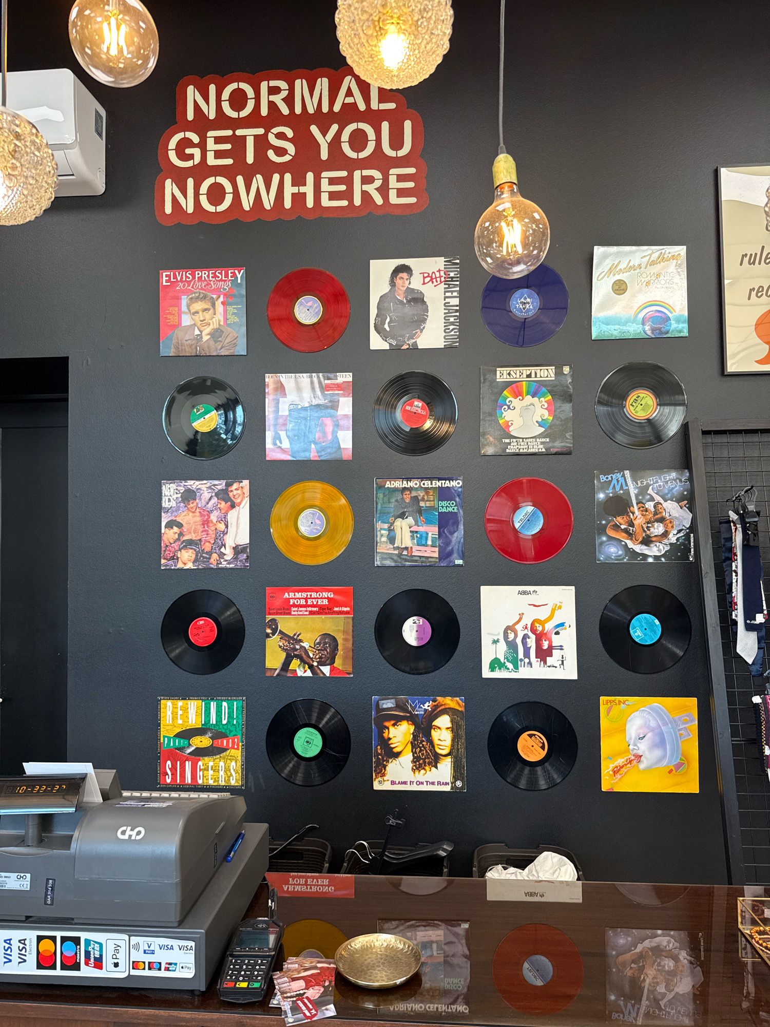 The image depicts the interior of a store, likely a record shop, with a black wall as the primary background. A large, red sign with white stencil lettering reads "NORMAL GETS YOU NOWHERE." The wall is adorned with numerous vinyl records and album covers, arranged in a grid-like fashion.
Notable album covers include "Elvis Presley: 20 Love Songs," a Michael Jackson album, "Modern Talking: Romantic Warriors," "Ekseption: The Little Dance," "Adriano Celentano: Disco Dance," "Armstrong for Ever," "Abba," "Boney M: Nightflight to Venus," "Blame It on the Rain," and "Lipps Inc." Several red and blue vinyl records are also displayed.
In the lower section of the image, a gray cash register is visible, with various payment card logos displayed on its side. A credit card terminal is situated to the right of the cash register, along with some small cards and a golden decorative plate. A metal rack with several ties hanging on it is partially visible on the far right. Three large, spherical light fixtures with visible incandescent bulbs hang from the ceiling, illuminating the scene. The overall impression is that of a vintage or retro-themed retail space.