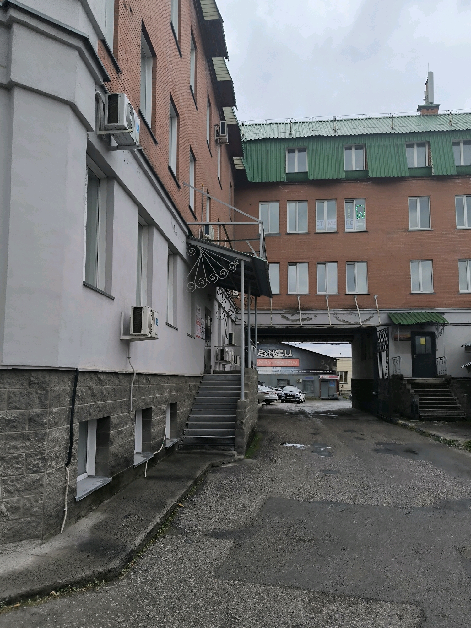 An overcast, mid-day exterior shot captures a narrow asphalt courtyard between two brick buildings in Novosibirsk, Russia. The building on the left, clad in grey stone and white stucco, features several large windows and an air conditioning unit mounted on its facade. A concrete ramp leads from the courtyard towards the building. The building on the right, constructed of red brick, has a green corrugated metal roof and numerous windows. A covered walkway, supported by metal beams, connects the two structures, forming an archway over the courtyard. Beneath the archway, two cars are visible parked on the asphalt. Stairs lead up to an entrance on the right side of the courtyard, with a closed dark door and a small green canopy overhead. The general impression is one of a functional, slightly utilitarian urban environment.