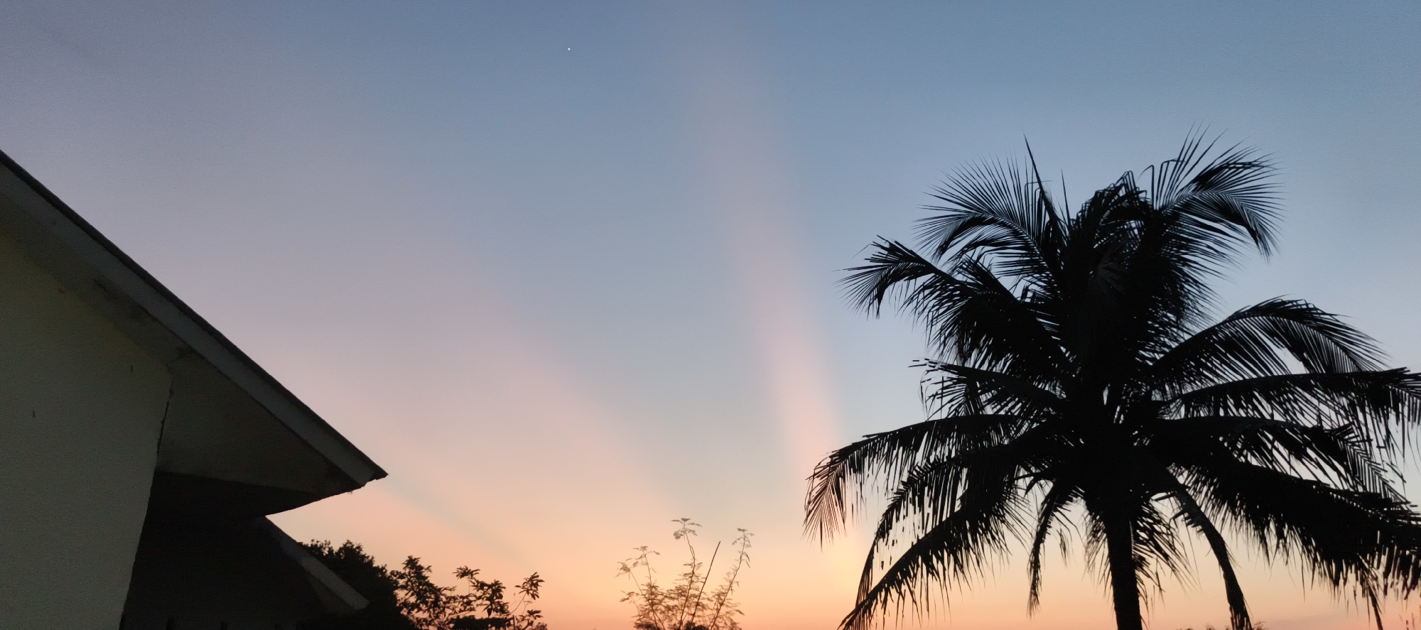 This is a horizontal, medium-shot photograph of a twilight sky with a silhouette of a palm tree and a portion of a building against the pastel hues of dusk. The sky transitions from a pale blue at the top to a soft orange and pink gradient towards the horizon. Faint crepuscular rays are visible, fanning out from an unseen point. A single, small, bright star or planet is visible in the upper center of the sky. On the left, the corner of a building with a cream-colored wall and a dark roofline is silhouetted. To the right, the dense silhouette of a palm tree with its fronds spread wide dominates the frame, with a few other smaller plant silhouettes visible at the bottom. The image captures a peaceful, tropical evening scene in Danau Lamo, Indonesia.