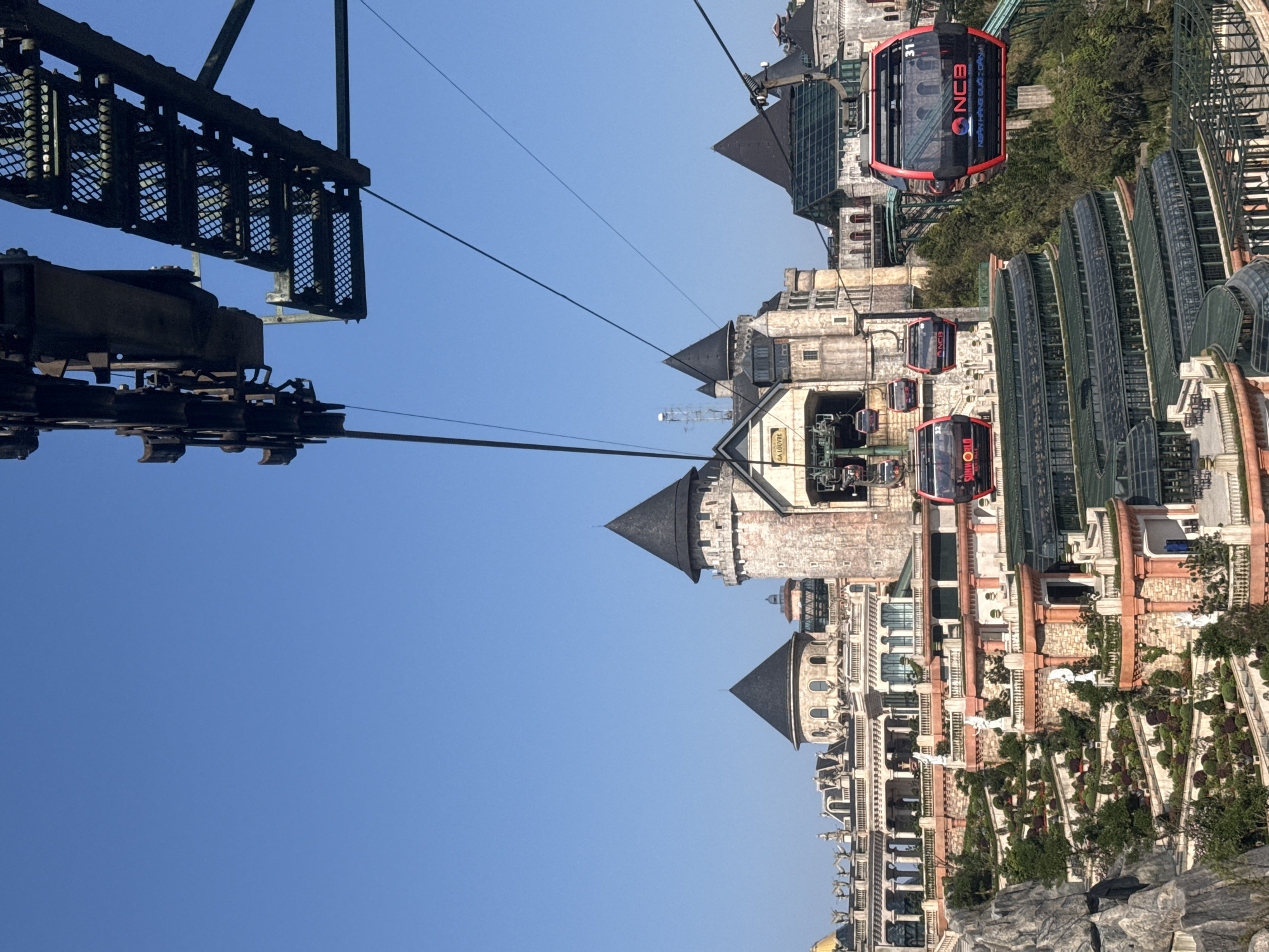 The image depicts an aerial perspective of Ba Na Hills, specifically the French Village area, located in the city of Đà Nẵng, Vietnam, under a clear blue sky.
In the upper portion of the frame, rusted metal components of a cable car system are visible, including large pulley wheels and a grated service platform. Multiple black cables extend diagonally downwards from these structures.
Several red and black cable car gondolas are present across the midground. One gondola, prominently in the right midground, bears the number "31" and the logo "NCB NGÂN HÀNG QUỐC DÂN". Further left, additional gondolas are seen near a station labeled "GA LOUVRE", with some marked "SUNWORLD". The gondolas are suspended from the cables, suggesting active operation.
Below the cable car lines, a sprawling complex of European-style architecture is visible, characterized by multi-story buildings with stone-textured facades, numerous windows, and distinctive dark conical roofs and spires. One structure on the left features a large golden dome. Interspersed within and around the buildings are extensive terraced gardens, meticulously landscaped with manicured shrubs and hedges, and adorned with white classical statues. Green, glass-roofed structures are visible in the lower midground. Rocky formations and dense green foliage are also present on the left and throughout the scene.
No individual people are clearly discernible within the frame.
