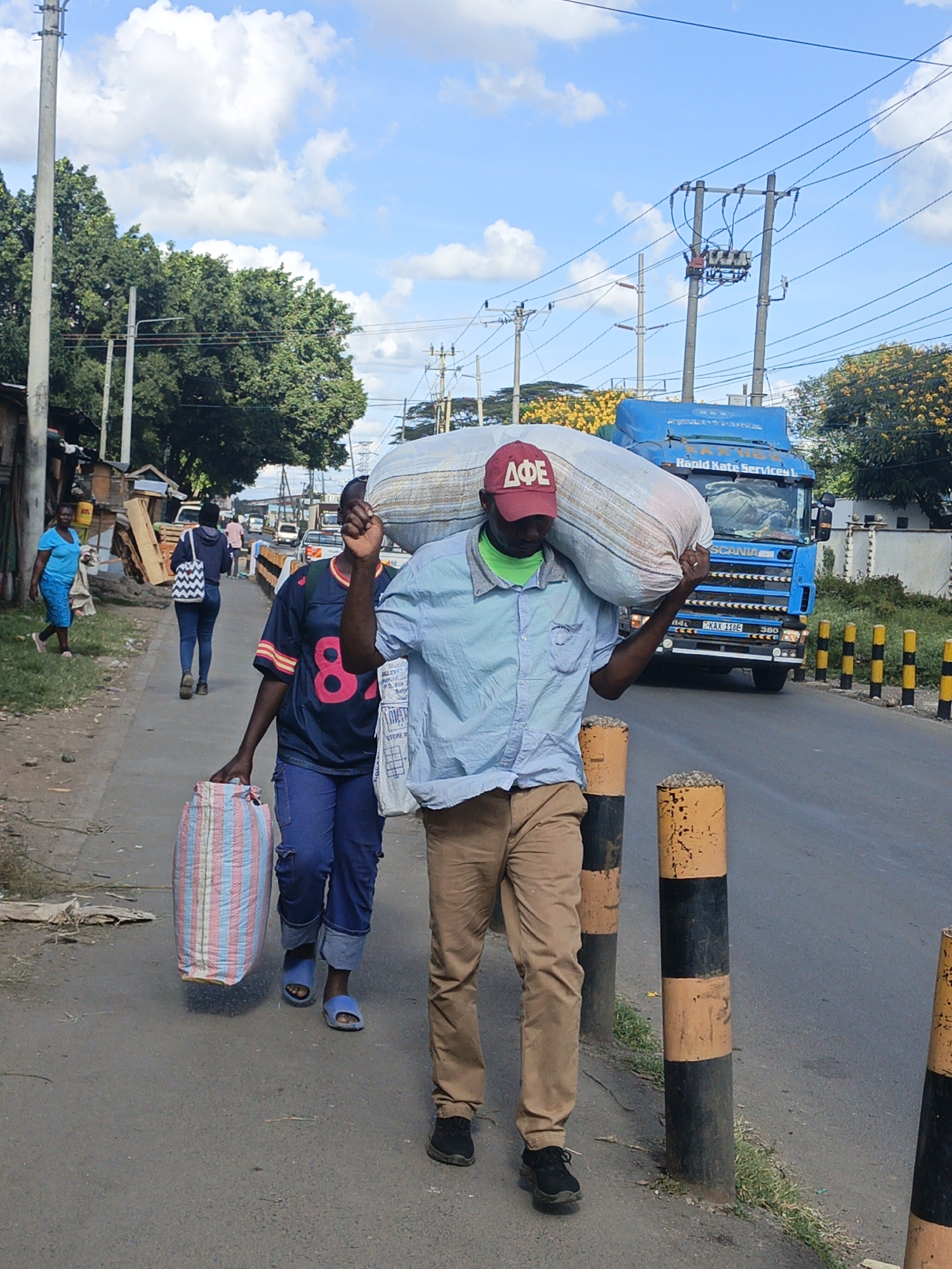 The image depicts a street scene in Nairobi, Kenya. A man wearing a red "ΔΦΕ" baseball cap, a light blue shirt, green neck gaiter, and khaki pants is carrying a large white sack on his shoulder as he walks along a paved road. Beside him on the sidewalk, another person, wearing a blue and red jersey with the number "84" on the back, blue pants, and sandals, carries a striped bag. Several other pedestrians are walking further ahead.
A blue truck with "Rapid Kate Services" written on it is visible on the road, partially obstructed by the man carrying the sack. Black and yellow bollards line the edge of the road. In the background, there are trees, buildings, utility poles with power lines, and other vehicles. The sky is blue with scattered clouds.