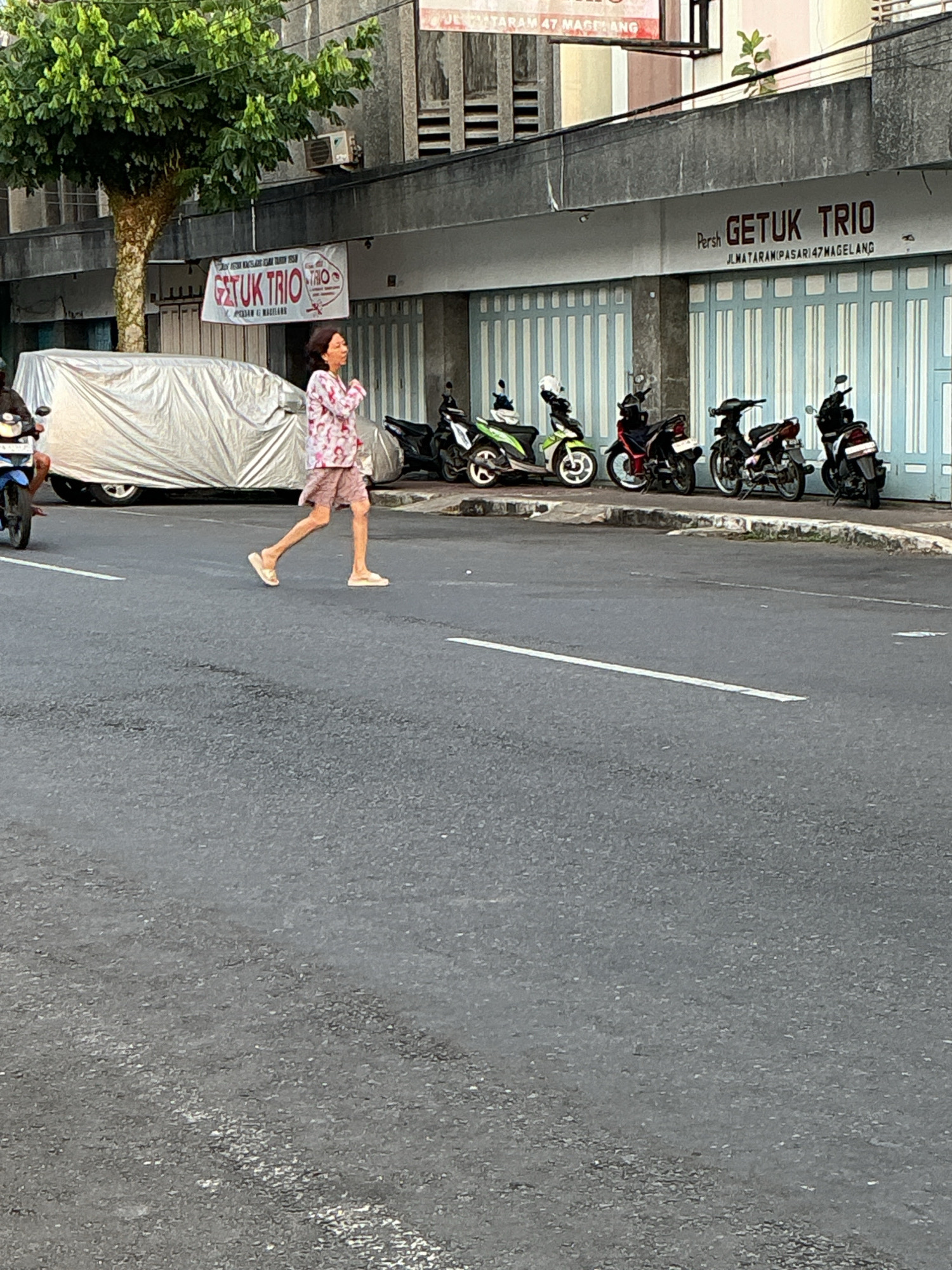 The image depicts a street scene in Magelang, Indonesia, likely in front of a business. A person is crossing the street, mid-stride, from left to right. They are wearing a light-colored, patterned shirt and shorts, and flip-flops. To the left of the pedestrian, a car is covered with a large, light-colored tarp. Behind the car and pedestrian, a building facade is visible with a sign that reads "GETUK TRIO" in dark lettering, with "JL.MATARAM (PASAR) 47 MAGELANG" underneath. The building has large, light blue and white striped doors. In front of the building, several motorcycles are parked along the sidewalk. A portion of a mature tree with green foliage is visible in the upper left quadrant of the image. The street itself is asphalt with a white line marking a lane. The overall environment suggests an urban setting during daylight hours.