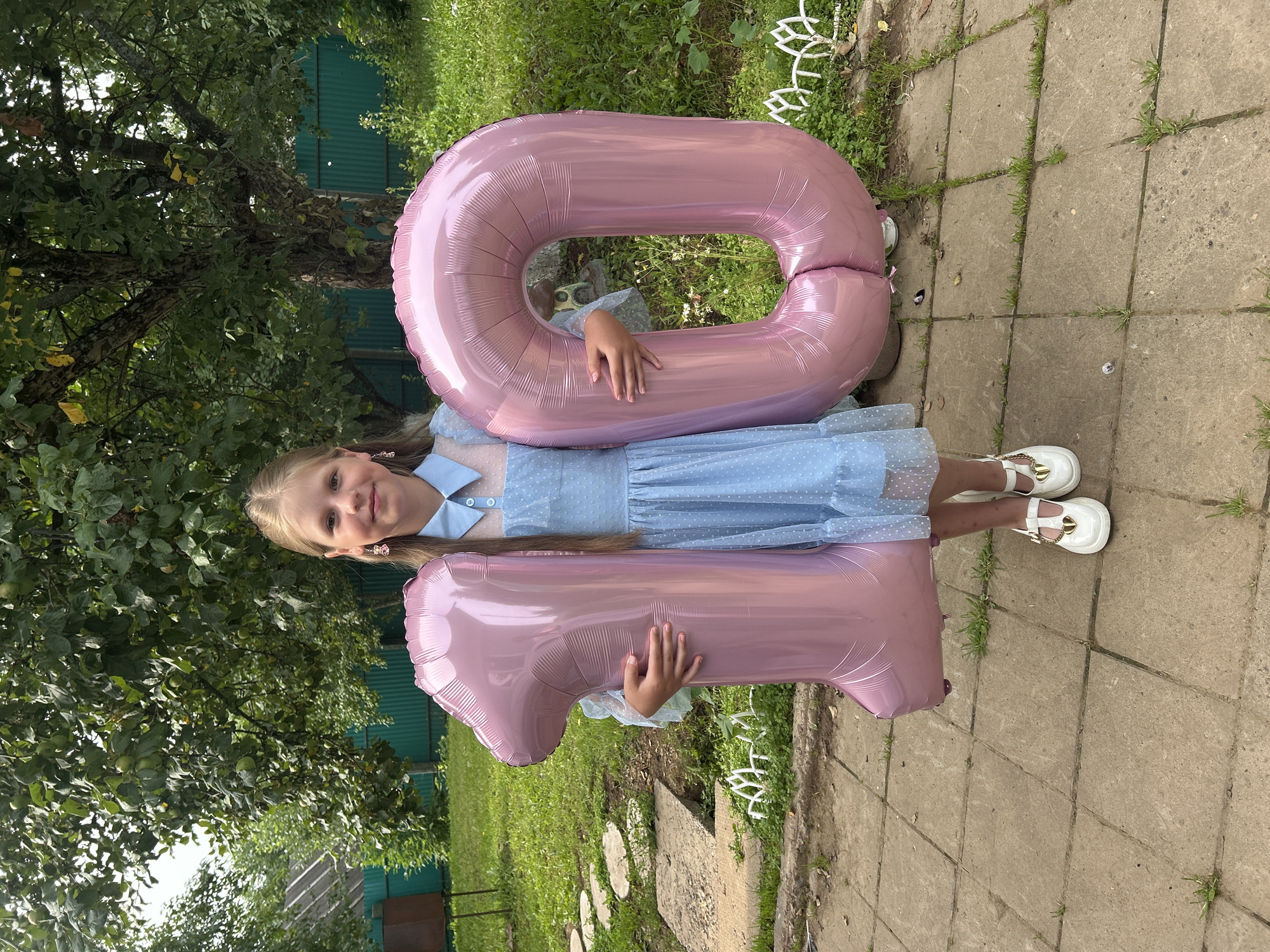 A young girl stands outdoors in a yard, holding two large pink balloons shaped like the numbers "1" and "0". She is wearing a light blue dress with white polka dots and white shoes with gold heart embellishments. The background shows a tree with green leaves and fruit, a green fence, and a stone pathway. The setting appears to be a residential yard. The image was taken in the city of Fokinskoye, Tver Oblast, Russia.