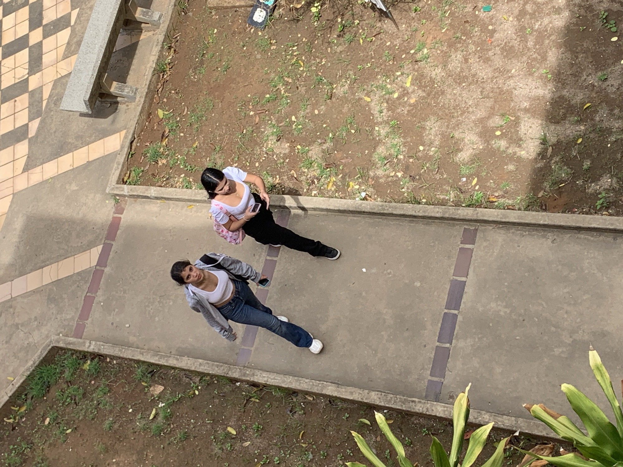Two young women are walking on a concrete path in a park-like setting. The woman on the left is wearing a grey zip-up hoodie over a white crop top and blue jeans. She has dark hair pulled back and is wearing white sneakers. She is holding a cell phone in her right hand. The woman on the right is wearing a white short-sleeved shirt with a pink patterned backpack. She has dark hair and is wearing black pants and black sneakers. She is looking down at her cell phone in her left hand. The path is bordered by dirt ground with sparse green vegetation and some fallen leaves. In the background, there is a checkered tiled area and a concrete bench. The scene appears to be in a public outdoor space, possibly a university campus or a recreational area.