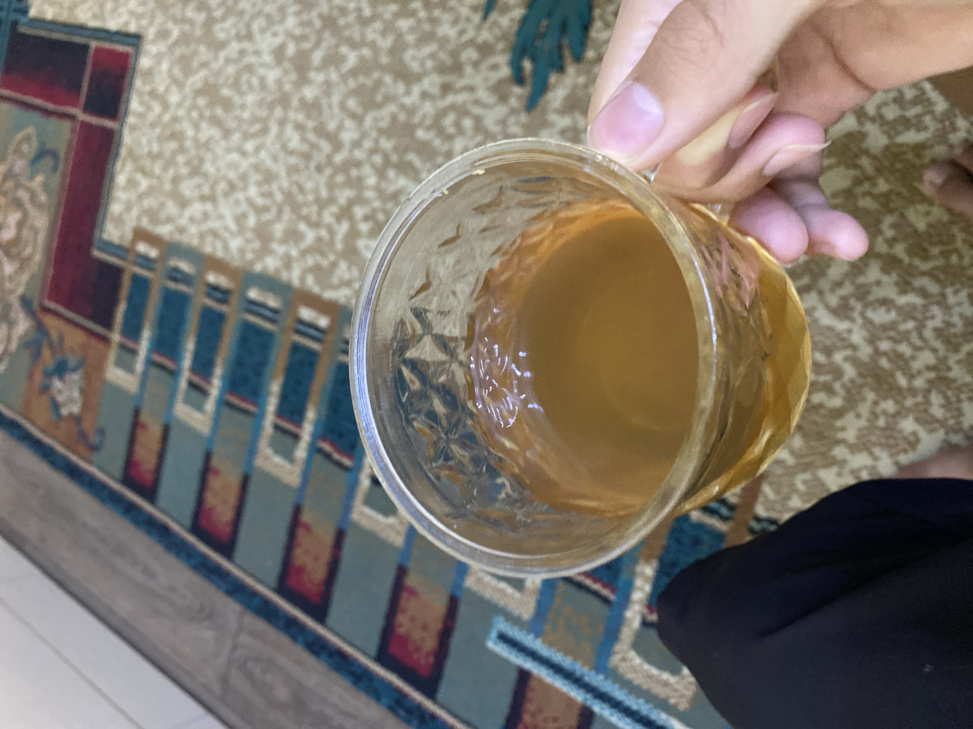 A person's hand holds a clear glass mug filled with a light brown liquid, likely tea. The mug has a textured surface. The background consists of a patterned carpet in shades of beige and a rug with geometric patterns in blue, red, and tan. The flooring adjacent to the carpet appears to be wood. The image is taken from a high angle, focusing on the mug and the hand. There is no discernible information indicating the location within the city of Turkistan, Kazakhstan.