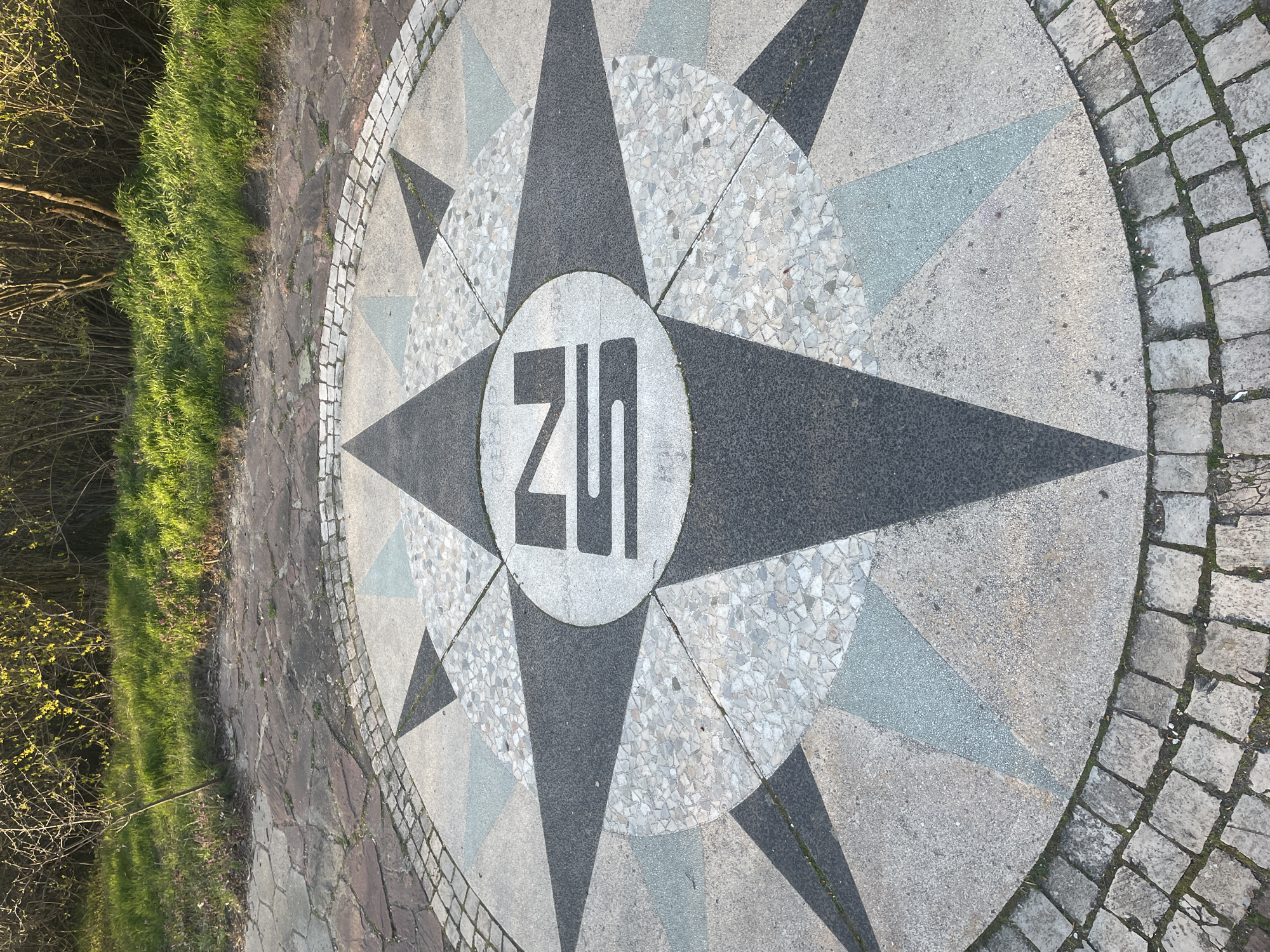 The image shows a compass rose inlaid in pavement, likely stone or concrete. The design features a central circle with the Cyrillic letters "ZU," surrounded by alternating dark and light-colored star points. The compass rose is bordered by cobblestones. Adjacent to the pavement is a grassy area and some foliage, suggesting an outdoor location.
This is likely the compass rose located at the site of the foundation stone of the National Palace of Culture (NDK) in Sofia, Bulgaria. The "ZU" likely stands for "ЗУ" (Zemlen Utvardzhdavan), which translates to "foundation" or "laying of the foundation".