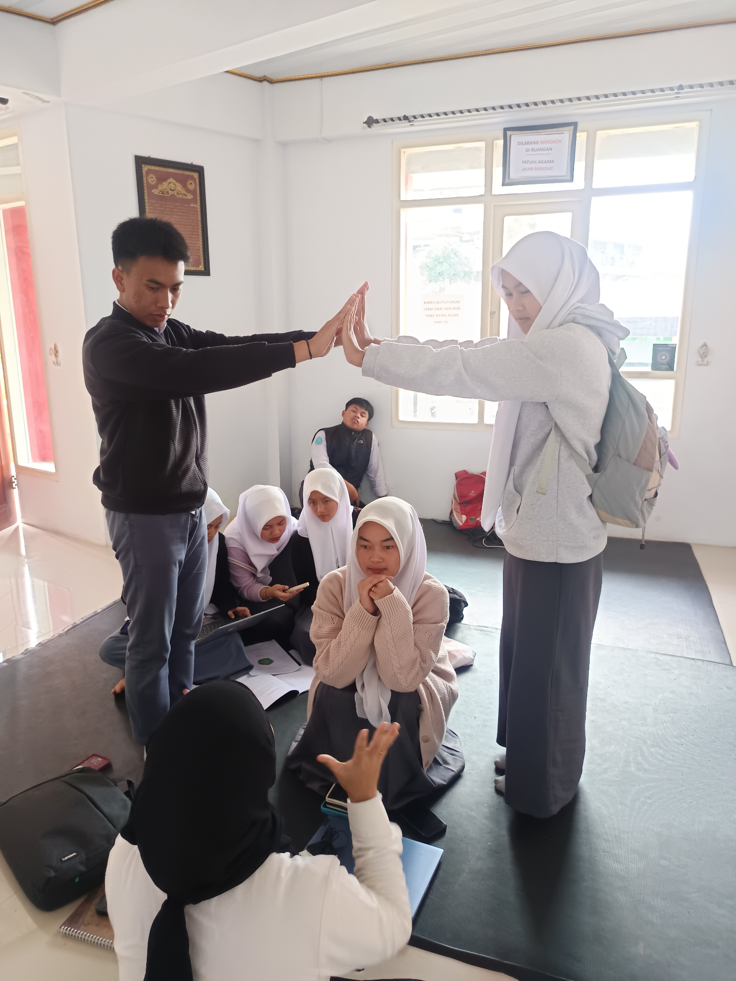 The image depicts a group of individuals indoors, likely in Gunung Putri, Indonesia, given the text on the sign. A young man and a young woman wearing a hijab are standing and raising their hands to touch palms, an interaction involving two people standing in front of them, several more are sitting on a dark mat in front of a window. They are surrounded by other young people who are seated on the floor, some of whom are using laptops or phones. There are also bags and papers scattered on the mat. A sign on the wall indicates a no-smoking policy and reminds people to adhere to religious principles. The setting appears to be a classroom or community space.