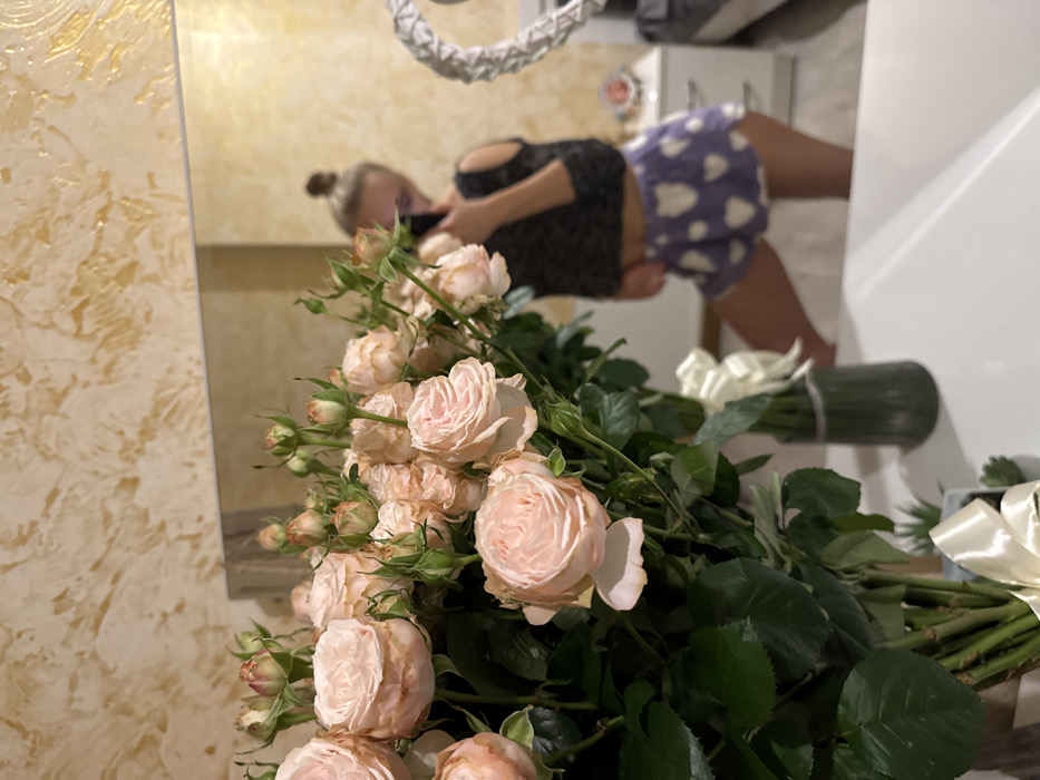 A bouquet of light pink roses with green leaves dominates the foreground. The roses are in various stages of bloom. Behind the flowers is a mirror reflecting a woman with her hair in a bun, wearing a black top and patterned shorts. The reflection also shows a white door or cabinet. A decorative white object hangs above the woman in the mirror. The scene appears to be indoors, possibly in a bathroom.