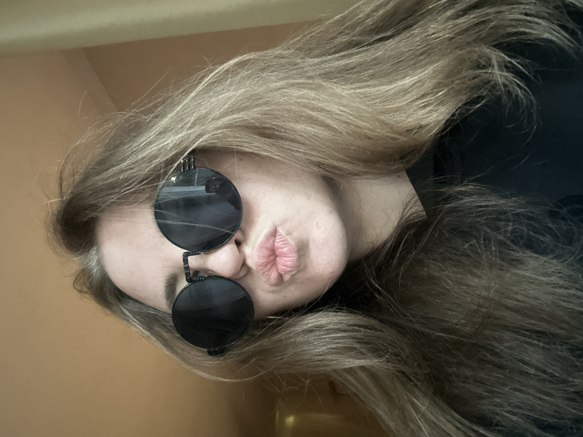 A close-up portrait captures a person, likely female, from the shoulders up, posing with a tilted head and a playful expression. The person has long, flowing blonde or light brown hair that appears somewhat disheveled, framing their face and cascading to the right side of the frame. They are wearing distinctive round, dark-lensed sunglasses with a black frame, which obscure their eyes. Their lips are pursed into a prominent pout or kissing face, revealing their pink, full lips.
The background on the left and top consists of a plain, light orange or tan wall, suggesting an indoor setting. To the right of the person's head and hair, a dark, indistinct area is visible, possibly part of their clothing or another background element. The lighting is soft and even, highlighting the texture of the hair and the details of the lips and sunglasses. The image conveys a casual, lighthearted mood. There is no visible text.
