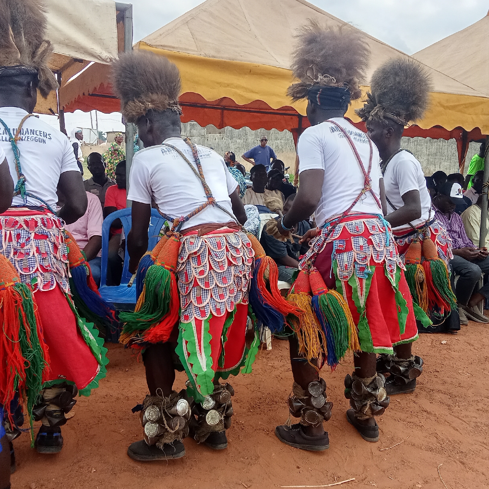 The image shows four men in Akwanga, Nigeria, viewed from behind, participating in a cultural dance. They wear white t-shirts with writing on the back, red and green skirts, elaborate headpieces made of fur or similar material, and ankle rattles made of what appears to be tin cans. The dancers are performing on a red dirt surface, with a crowd of spectators visible in the background. There is a tent-like structure with a yellow and red awning behind them.