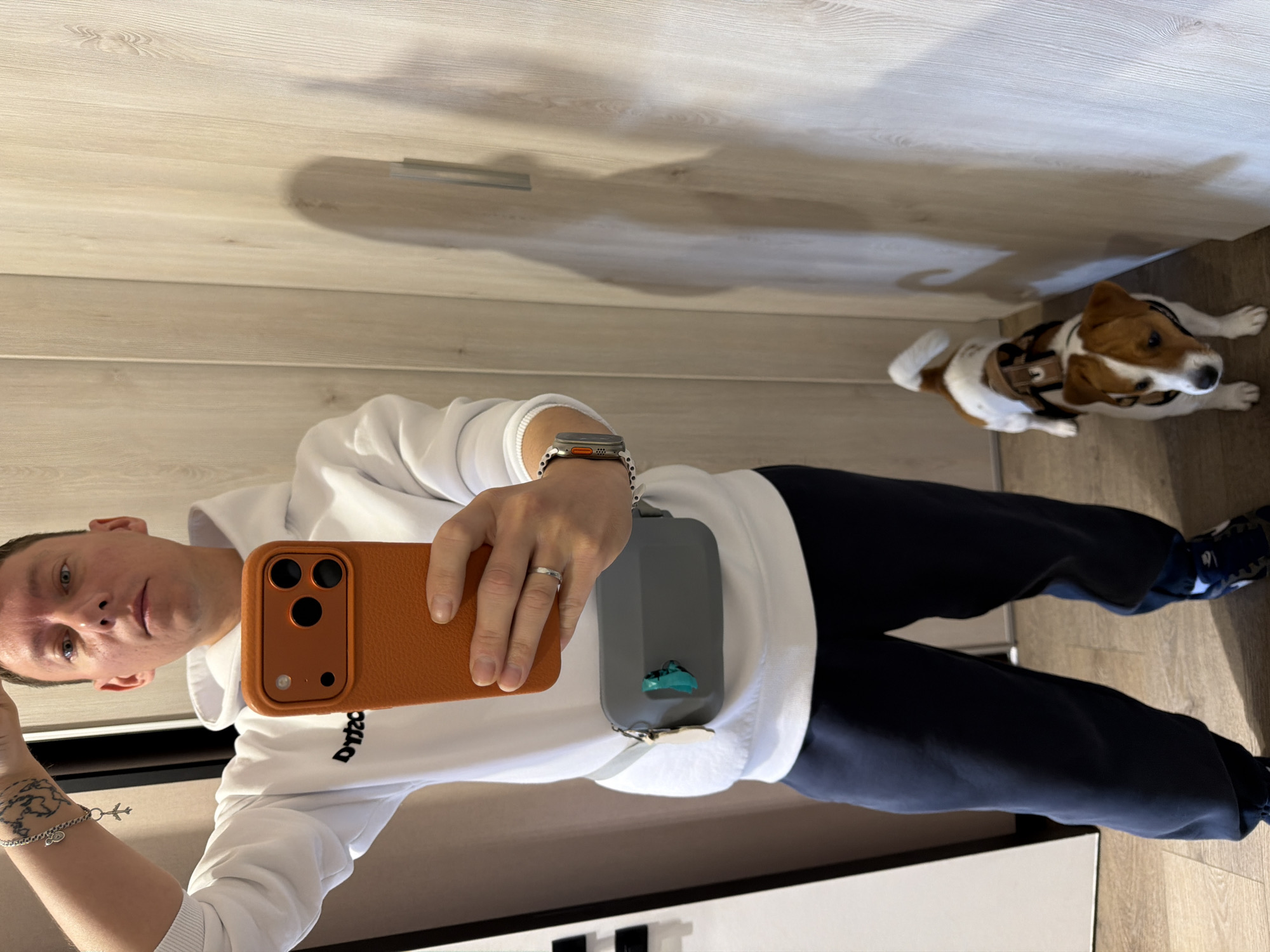 A man and his dog stand in a hallway with wood-paneled walls. The man is wearing a white hoodie, dark blue sweatpants, and a smartwatch. He is holding an orange smartphone and has a gray fanny pack around his waist. The dog, a Jack Russell Terrier, is wearing a harness and looking at the camera. The scene appears to be indoors, possibly in a residential building, as indicated by the hallway and wood paneling. There is no indication of the specific location within Moscow, Russia.