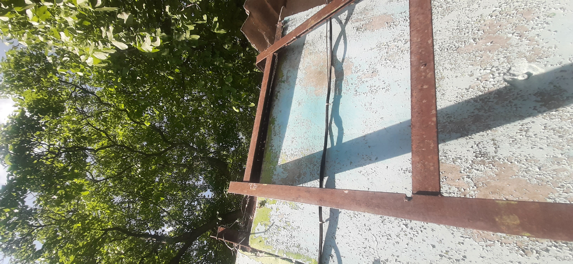 An upward angle shot captures a weathered, light blue structure with peeling paint, topped by a rusty metal frame. Above this structure, lush green tree foliage against a bright sky dominates the upper portion of the image. The scene appears to be outdoors, likely in a residential or industrial area, under clear daylight. There are no people or animals visible. The setting could be the city of Yessentukskaya, Russia, based on the provided information.