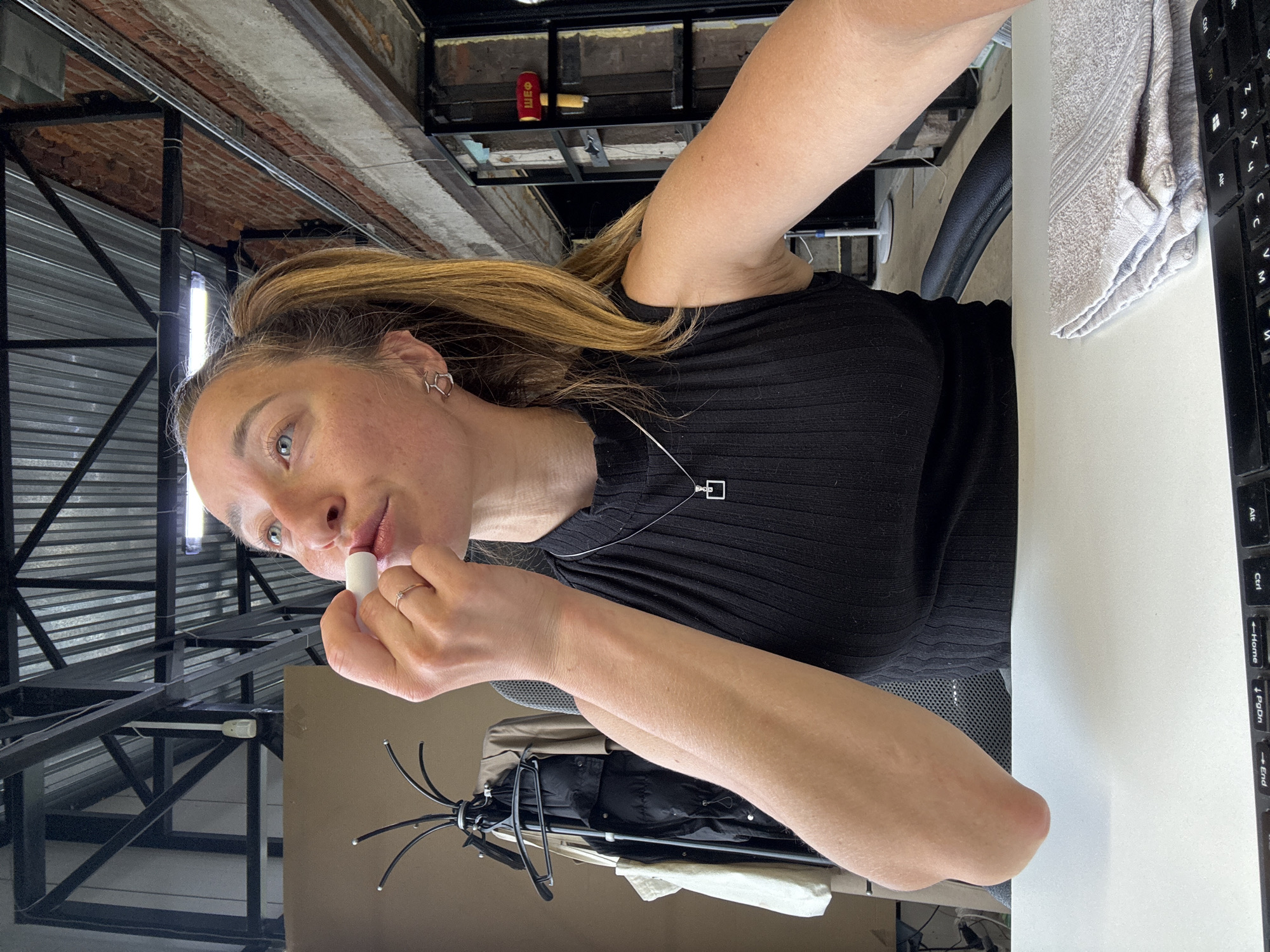 A woman is applying lip balm. She has blonde hair and blue eyes, wearing a black ribbed shirt and a delicate necklace. She is sitting at a desk with a keyboard and a towel. The location is indoors, potentially an office or studio, with a metal beam structure visible in the background. The city of Chaikovsky, Russia is not discernible from the image.