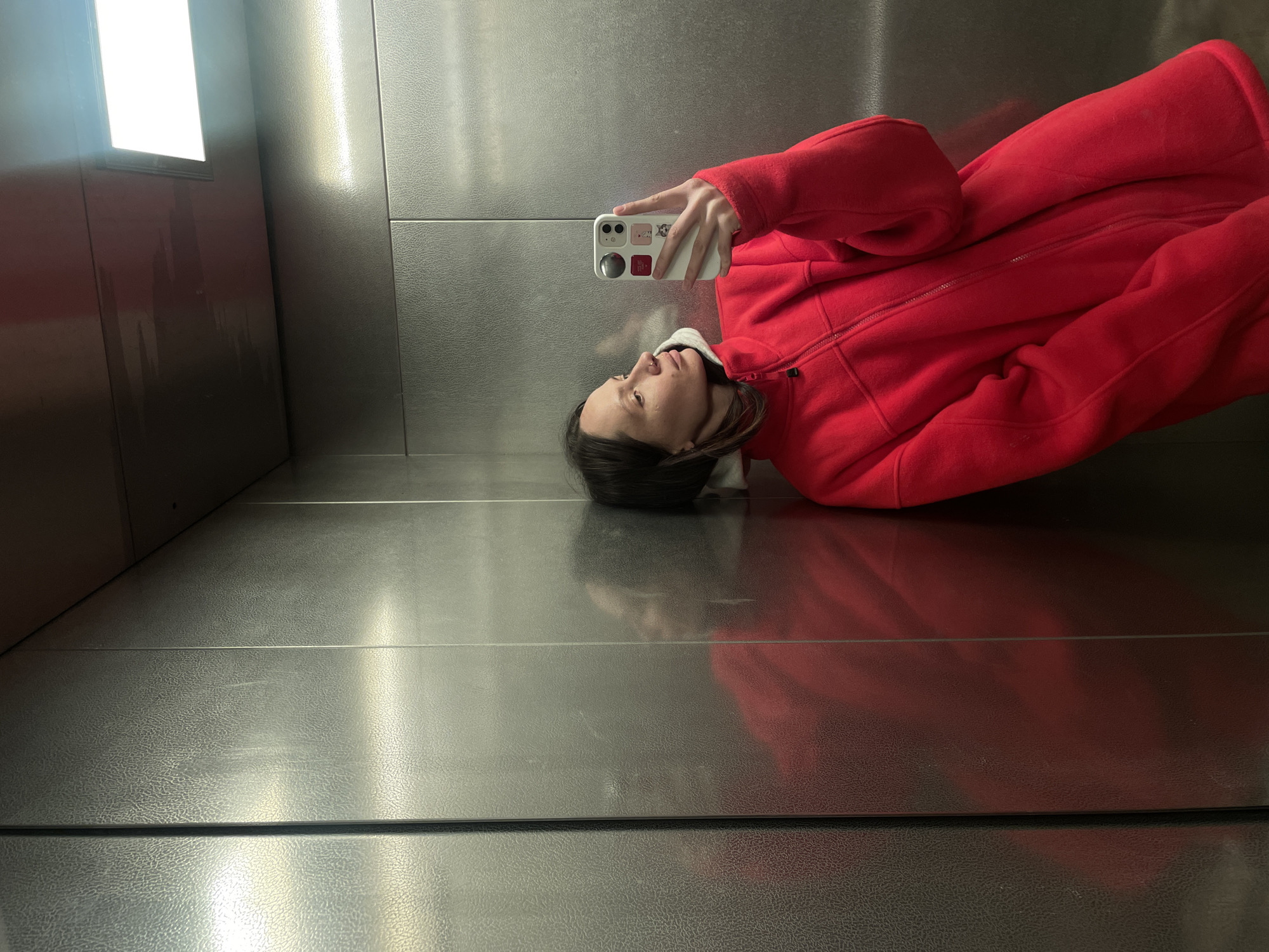 A person wearing a red zip-up fleece jacket is taking a selfie in what appears to be an elevator. The elevator has a gray, textured metal interior. The person is holding a white smartphone with stickers on the back of the case. The location is not identifiable as Izhevsk, Russia, based solely on the visual information.