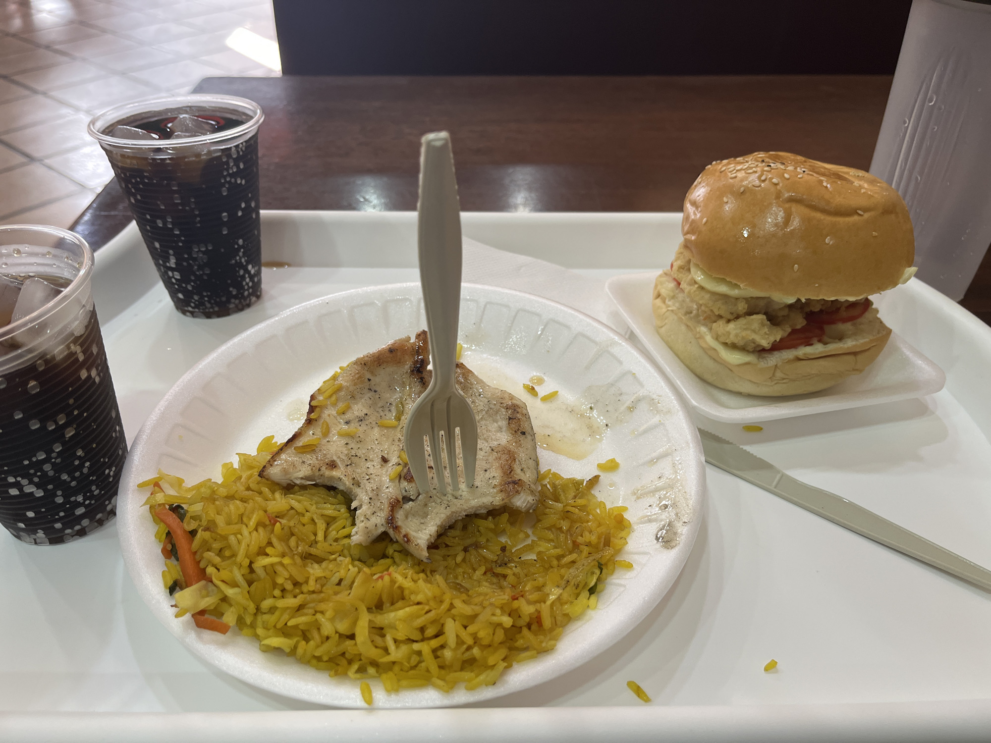 The image depicts a meal on a white cafeteria tray. The meal consists of yellow rice, a piece of grilled chicken pierced with a white plastic fork, a hamburger on a separate white plate, a white plastic knife, and two plastic cups filled with a dark beverage with ice. The scene is likely indoors, potentially in a food court or cafeteria. While it is claimed to be Porlamar, Venezuela, that cannot be verified from the image.