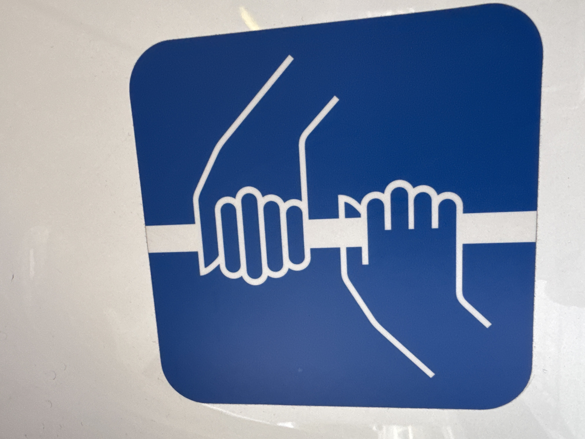 This is a close-up, eye-level, indoor shot of a blue and white graphic symbol against a light gray, reflective surface. The symbol is square with rounded corners and features a stylized representation of two hands holding a horizontal bar.
The left hand, depicted in white outline on the blue background, is gripping the bar with its fingers splayed. The thumb is not clearly visible, but the general shape suggests a firm grip. A white line extends upwards and to the left from the wrist area, representing the forearm or perhaps a connection to another part of the symbol.
The right hand, also in white outline, is positioned on the right side of the bar. Its fingers are also curled around the bar, and its thumb is visible, pointing downwards. Similar to the left hand, a white line extends upwards and to the right from the wrist area. The horizontal bar itself is depicted as a thick white line across the center of the symbol, connecting the two hands.
The symbol is clean and appears to be printed or applied onto the surface. The background surface shows some subtle reflections and slight variations in texture, indicating it is likely a smooth, man-made material. There is no visible text in the image. The overall impression is that of a safety or instructional symbol, likely indicating a point of leverage, support, or an area that requires a two-handed grip.