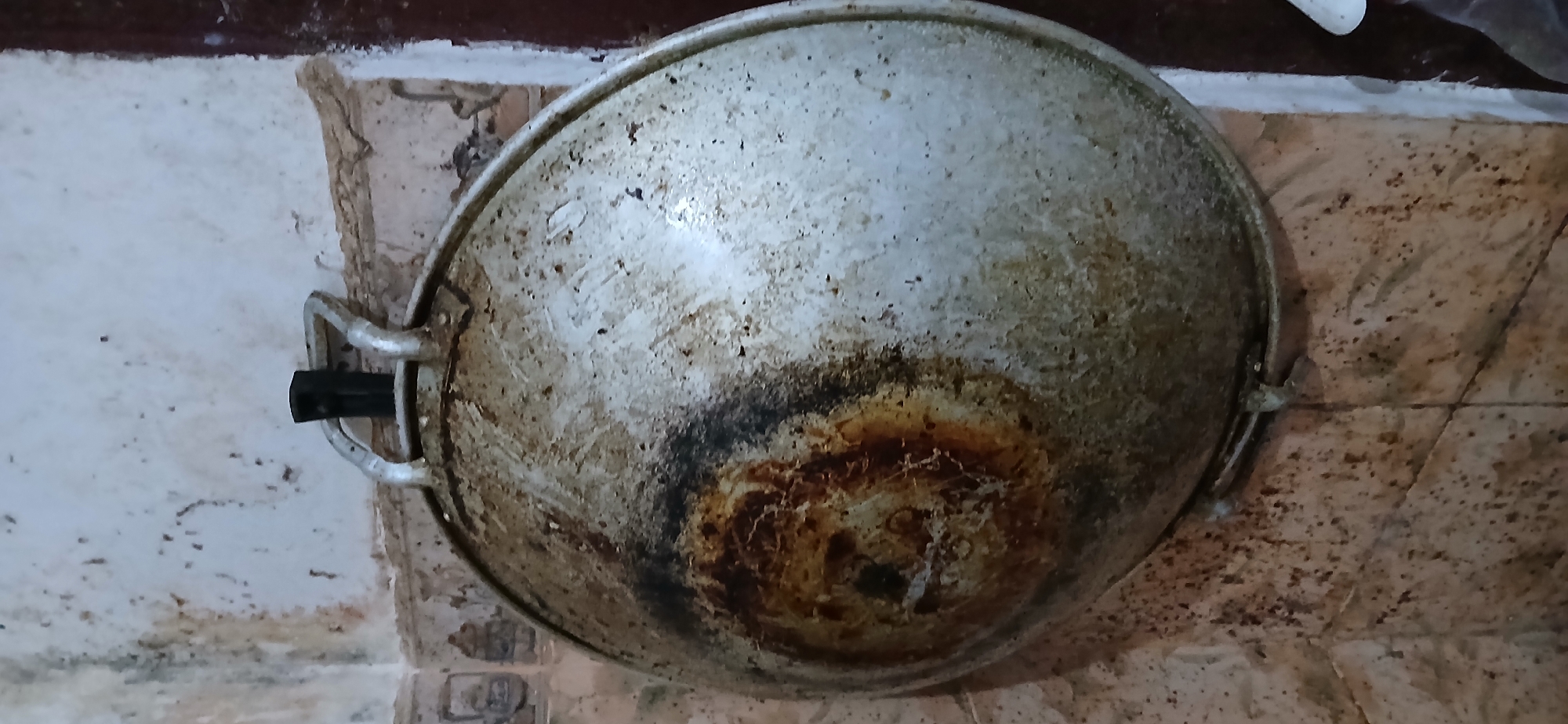 A well-used, large, round, metal cooking pan hangs by its handles from a hook on a textured, off-white, discolored wall. The pan exhibits significant discoloration and burn marks, particularly on the left side, indicating past cooking activity. The wall appears to be tiled with a light-colored, possibly marble or ceramic, pattern that is also dirty and stained. The overall environment suggests a kitchen or cooking area that is not meticulously maintained. The specific location in Samarinda, Indonesia, is not discernible from the visual information provided in the image.