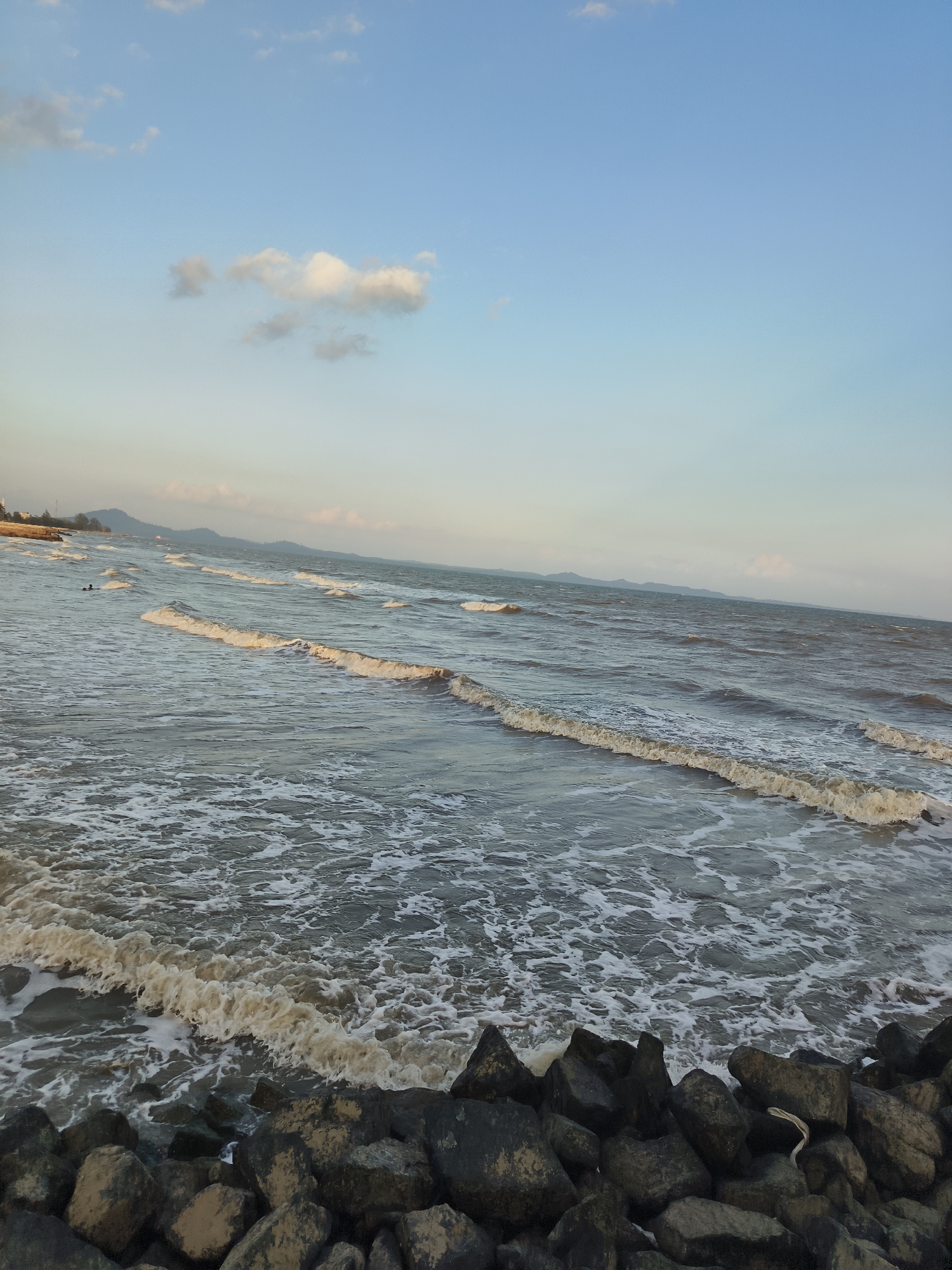 The image depicts a coastal scene. The foreground is dominated by a pile of dark, irregularly shaped rocks. Beyond the rocks, the ocean stretches towards the horizon, with waves breaking and creating white foam. The water appears murky brown. In the distance, the shoreline is visible, featuring a line of trees and some structures, possibly buildings, suggesting a developed area. Further in the background, hills or mountains are faintly visible under a clear sky with a few scattered, wispy clouds. The sky is a gradient of light blue, transitioning to a paler hue near the horizon. The overall impression is of a somewhat overcast day with calm seas. It is not possible to determine the specific location in the city of Kupang Berkah Jaya, Indonesia, from the visual information provided. There is a single indistinct human figure visible in the water, appearing to be swimming.