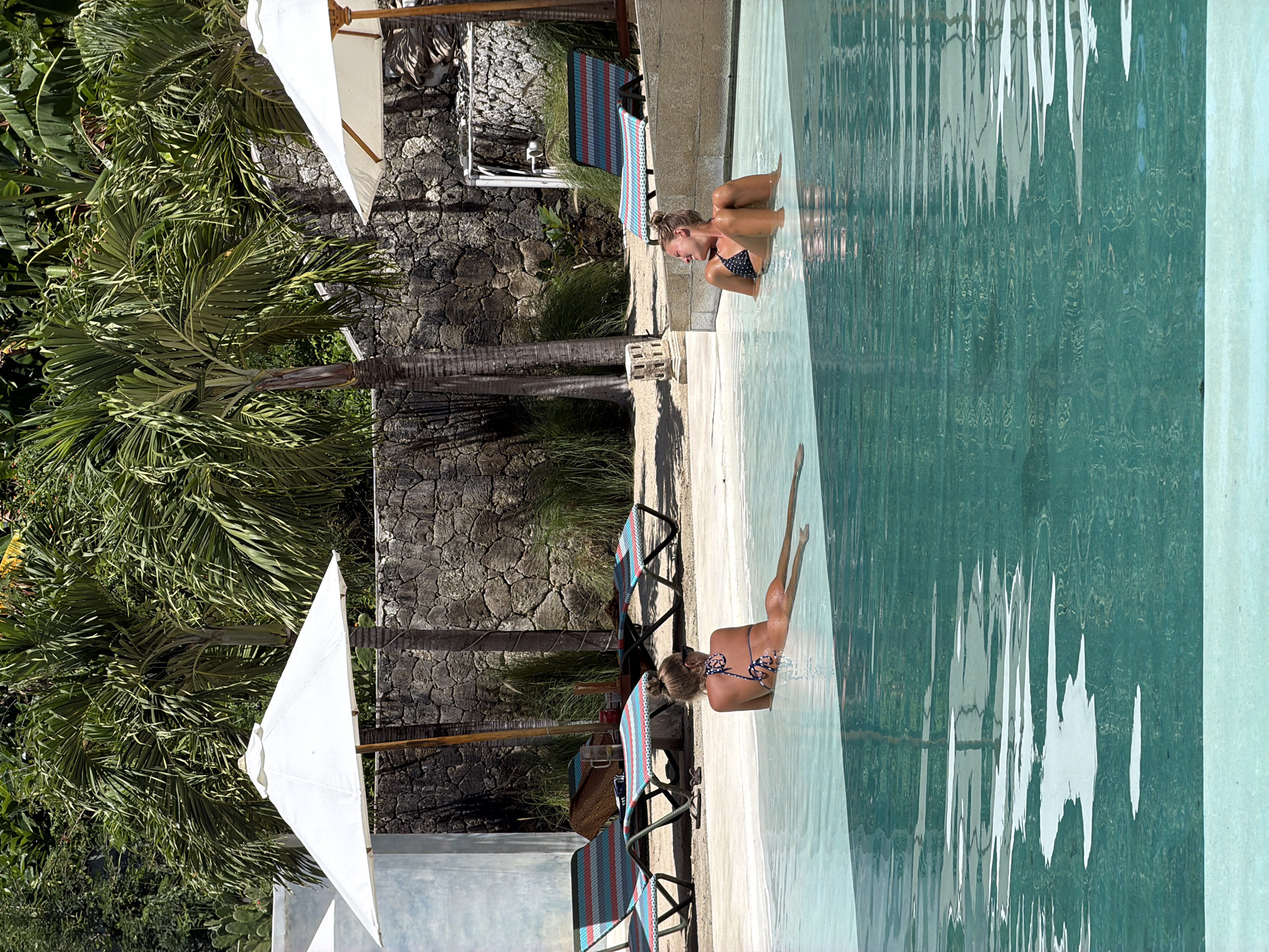 The image shows a swimming pool area in Batununggal, Indonesia. Two women in bikinis are at the edge of the pool. One woman is submerged up to her chest, while the other is in the water with her legs extended. Adjacent to the pool, there are beach chairs and white umbrellas. The background features a stone wall and palm trees. The water in the pool is greenish-blue, with surface reflections.