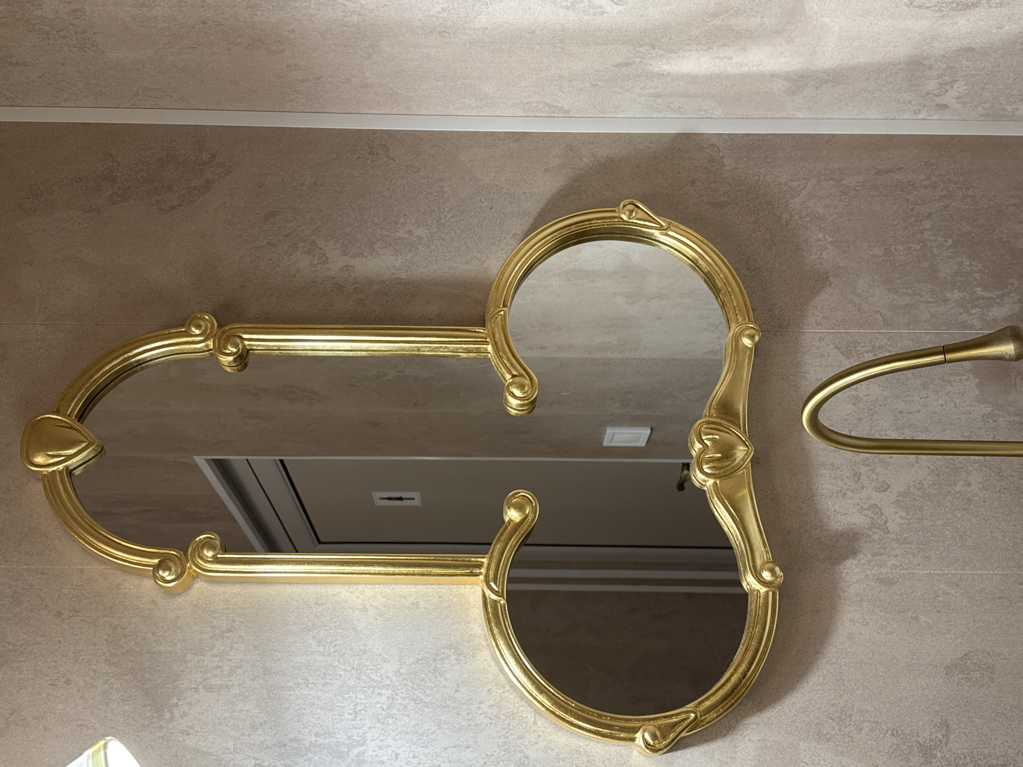 The image shows a gold-framed mirror mounted on a light brown tiled wall. The mirror has an unusual design, with two circular mirrors connected by a rectangular one. There is also a gold-colored towel rack visible on the right side of the mirror. The wall is decorated with a horizontal white strip near the top. The location is unknown.