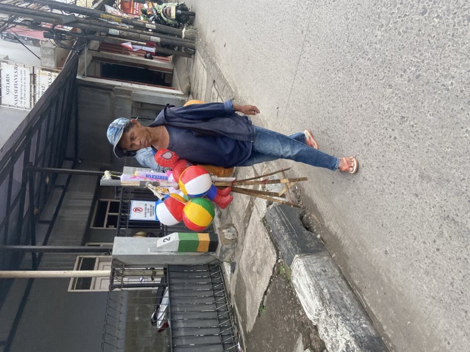 A man is walking down a paved street in Bojongsoang, Indonesia (latitude: -6.972, longitude: 107.636). He is carrying a stand with inflatable toys, including a Spiderman, a colorful ball, and a striped ball. He is wearing a blue shirt, blue jeans, a blue hat, and orange sandals. He is walking on a sidewalk next to a concrete curb and a black iron fence. On the opposite side of the street, there are buildings with a sign for a notary's office. There is a motorcycle parked on the side of the street.