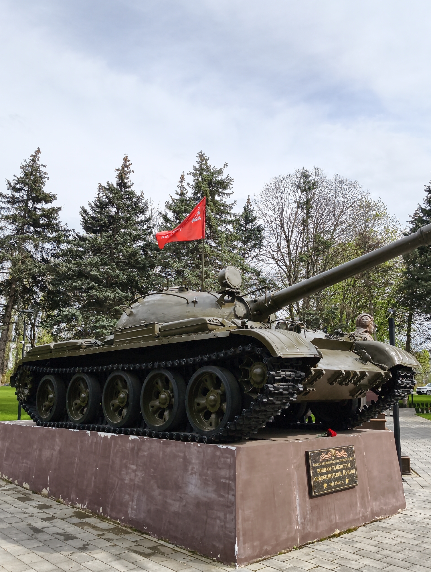 An olive-green Soviet-era tank, likely a T-54 or T-55, stands as a monument in Krasnodar, Russia. The tank is positioned on a rectangular, reddish-brown concrete pedestal, which shows some signs of wear and flaking paint. The tank's long main gun points to the right. It features prominent tracks and five large road wheels on each side. A red flag, emblazoned with a gold hammer and sickle and star, is mounted on the tank's turret, slightly unfurled by a breeze. A small bust or figure is visible protruding from an open hatch on the right side of the turret.
In front of the monument base, the ground is paved with light-colored square tiles. A small, dark rectangular plaque is affixed to the front of the pedestal, beneath which a single red rose is laid. The background is dominated by numerous tall, dense coniferous trees, interspersed with some bare deciduous trees, suggesting a park or memorial setting. The sky above is overcast and light grey, indicating daytime with diffused natural light.
The plaque on the monument reads:
"70 ЛЕТ
ПОБЕДА В ВЕЛИКОЙ ОТЕЧЕСТВЕННОЙ ВОЙНЕ
ВОИНАМ-ТАНКИСТАМ,
ОСВОБОДИТЕЛЯМ КУБАНИ
☆ 1941-1945 г.г."
This translates to:
"70 YEARS
VICTORY IN THE GREAT PATRIOTIC WAR
TO THE WARRIOR-TANKMEN,
LIBERATORS OF KUBAN
☆ 1941-1945"
The monument commemorates the 70th anniversary of the victory in the Great Patriotic War, honoring the tank crews who liberated the Kuban region. No active event is taking place; the scene depicts a static memorial display.