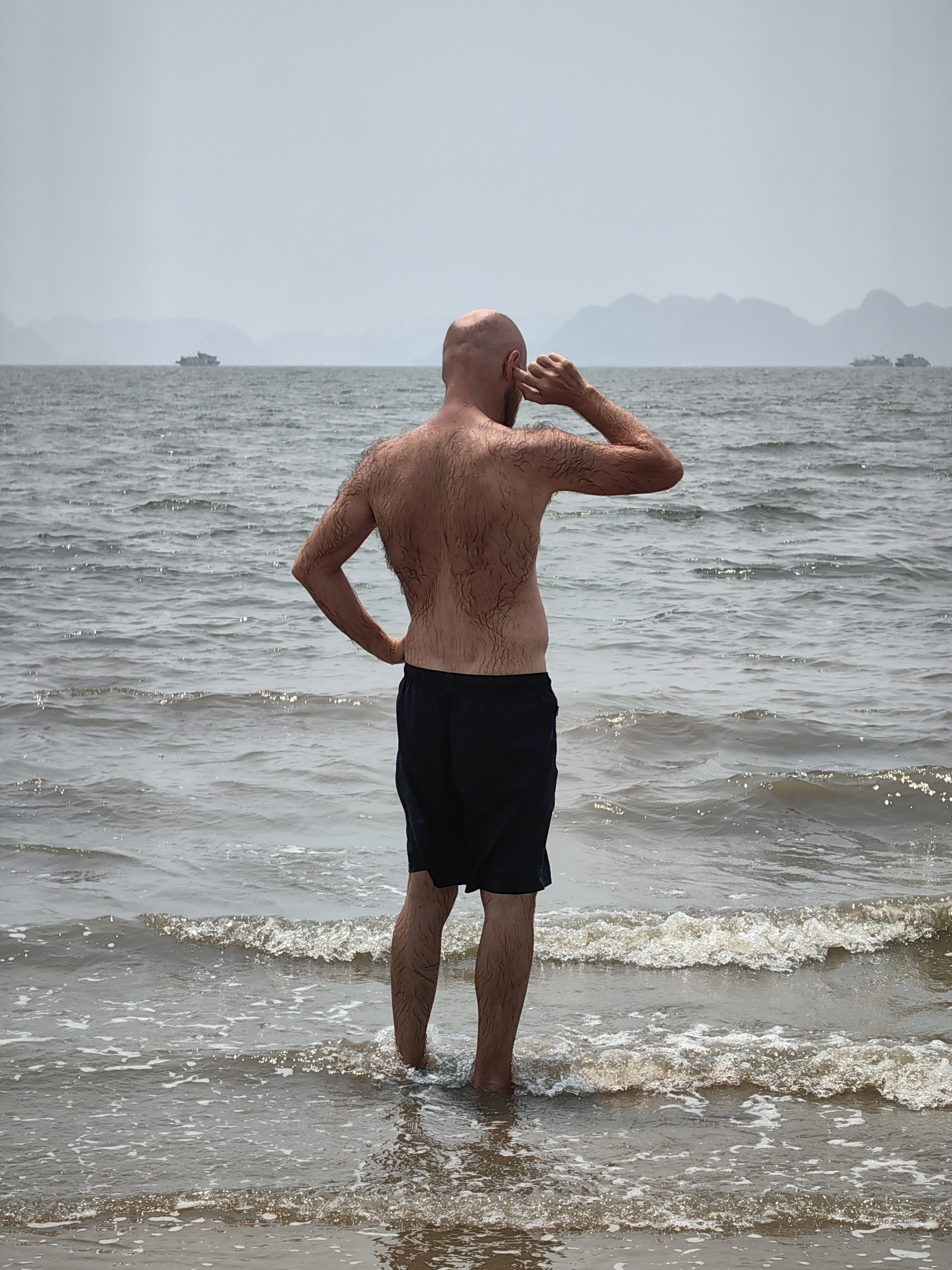 A bald, light-skinned man stands in the water at a beach in Bai Chay Ward, Vietnam. He is facing away from the viewer, with the ocean in front of him. The man is wearing dark blue swimming shorts. His right hand is on his hip and his left hand is near his ear. The water is at his lower calf level. In the distance, there are several ships on the water, as well as mountains in the background. The sky is overcast.