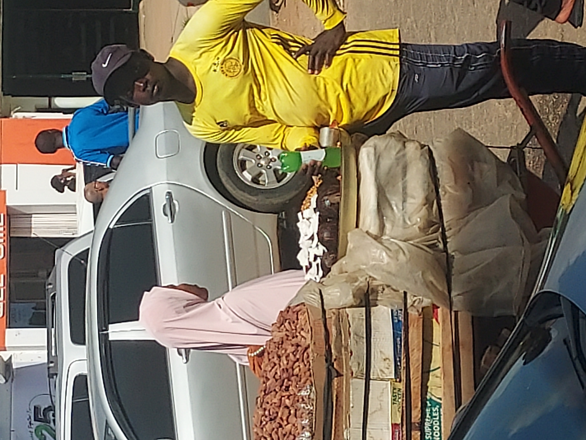 This image captures a vibrant street vending scene in Jos, Nigeria, during the daytime under bright, clear weather. The central figure is a male vendor wearing a bright yellow long-sleeved shirt with black stripes on the sleeves, dark trousers, and a dark cap with a white Nike logo. He stands behind a makeshift stall, looking directly at the camera.
His stall displays various items for sale: a large pile of roasted nuts or seeds, possibly groundnuts, in a wooden tray; several small white plastic cups; a green plastic bottle with a white pump or nozzle; and a clear plastic bottle, likely containing a beverage. Behind these items, stacks of cardboard boxes, some wrapped in white plastic bags, are visible. Text on the boxes includes "TASTY CHICKEN" and "SUPREME NOODLES," suggesting he also sells packaged food items.
In the background, a silver car is parked, partially obscuring another person in a blue shirt. Further back, near another car, a person in a light pink flowing garment is visible. The scene suggests a busy urban environment, typical of a street market or roadside vendor setup. The ground appears to be concrete or asphalt. A numerical "50" is also visible on a distant sign or package. The overall atmosphere appears to be a normal, bustling daily street activity.